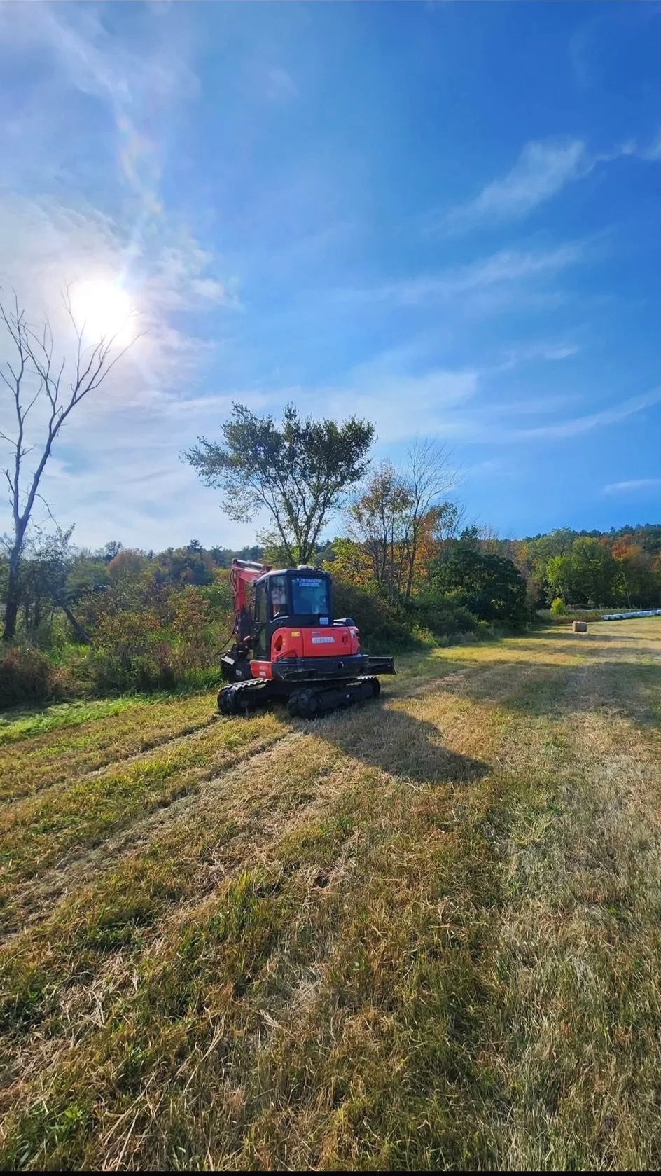 A red mini excavator parked on a grassy field with trees and a blue sky with some clouds in the background.