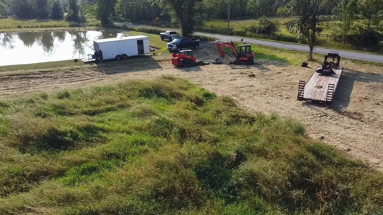 An outdoor construction scene by a pond with a trailer, two small excavators, a pickup truck, a flatbed trailer, and some work tools, surrounded by trees and grass.