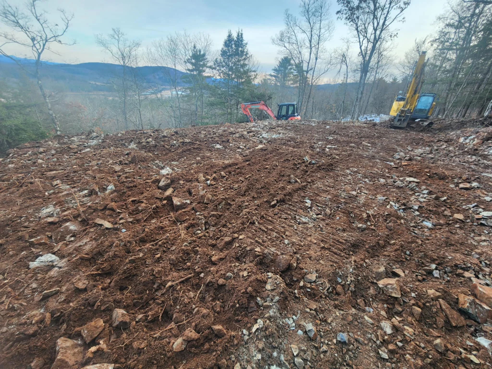 A construction site with excavators on a hillside, with trees and mountains in the background.