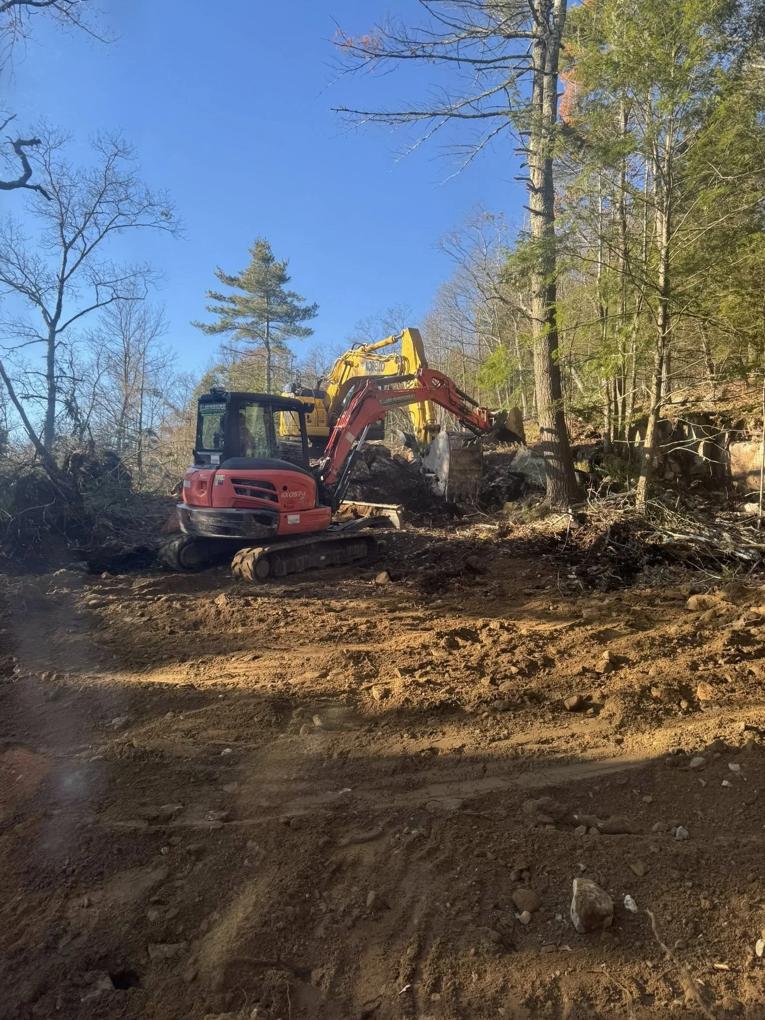 Construction site in a wooded area with dirt ground and trees, a small red and yellow excavator digging in the dirt.