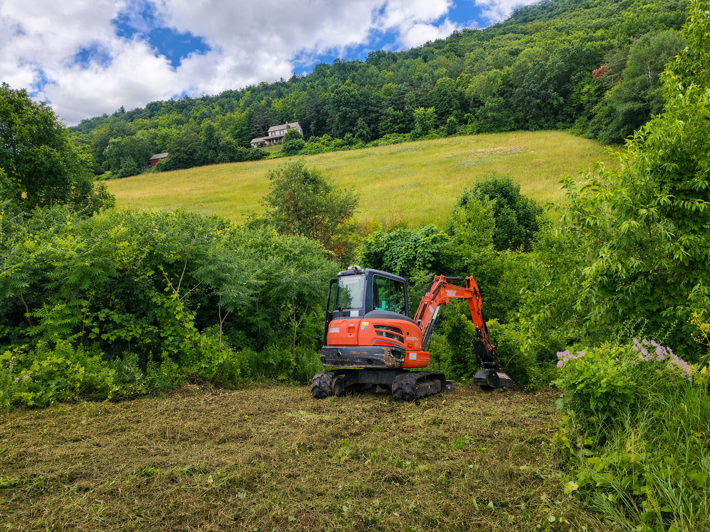 A small orange excavator working on a patch of cleared ground in a lush green hillside farm, with trees, bushes, and a house visible in the distance under partly cloudy sky.
