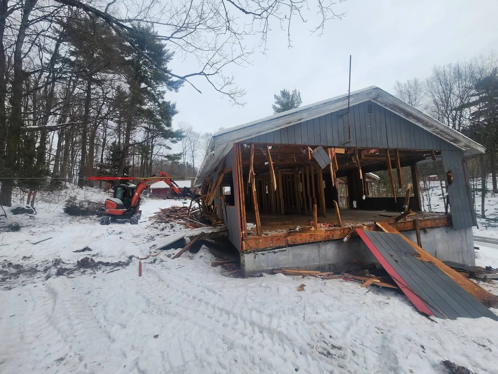 A partially demolished house in a snowy wooded area with construction equipment nearby.