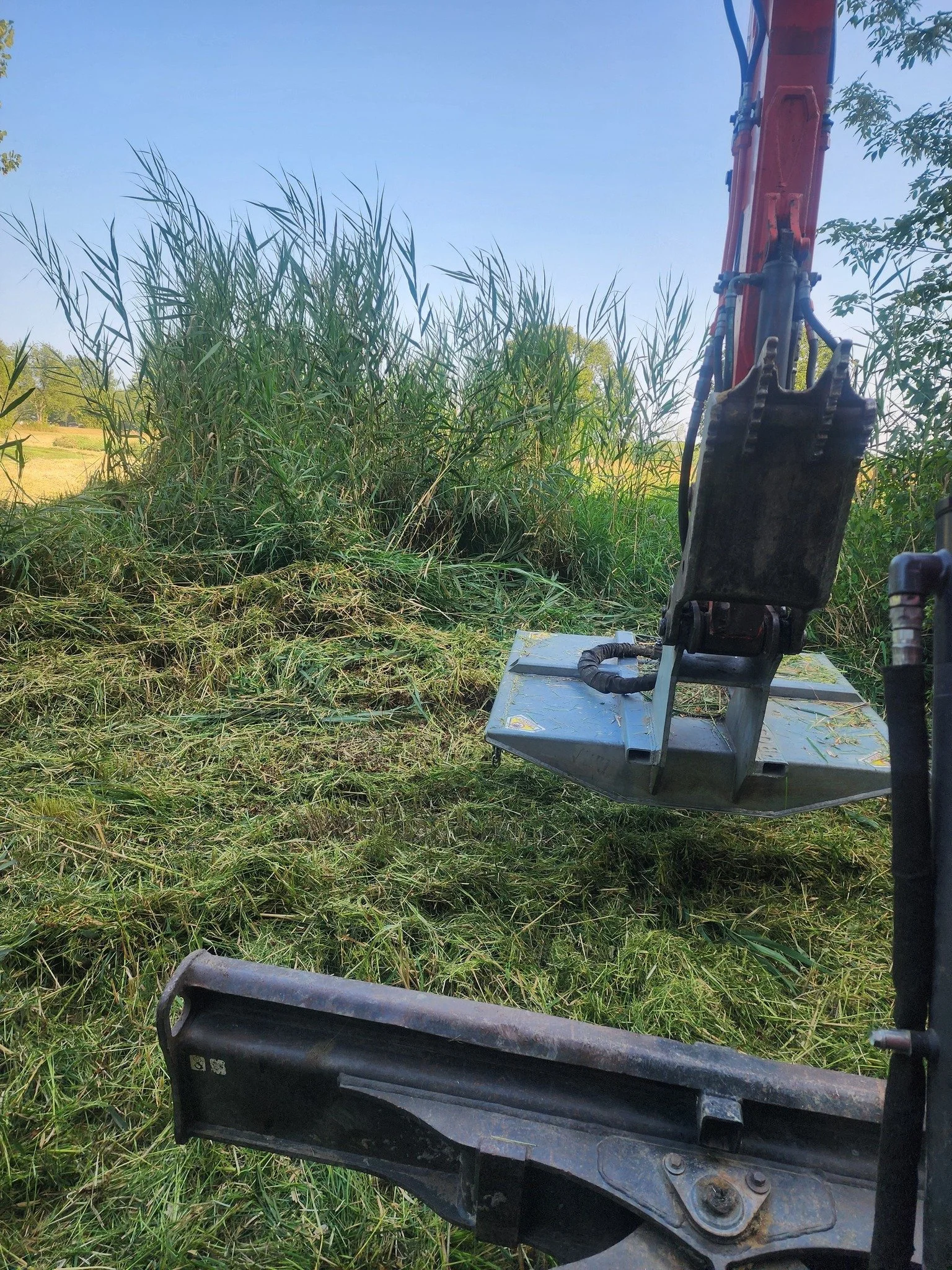 Close-up of excavator arm and bucket on a construction site, with grass and trees in the background.