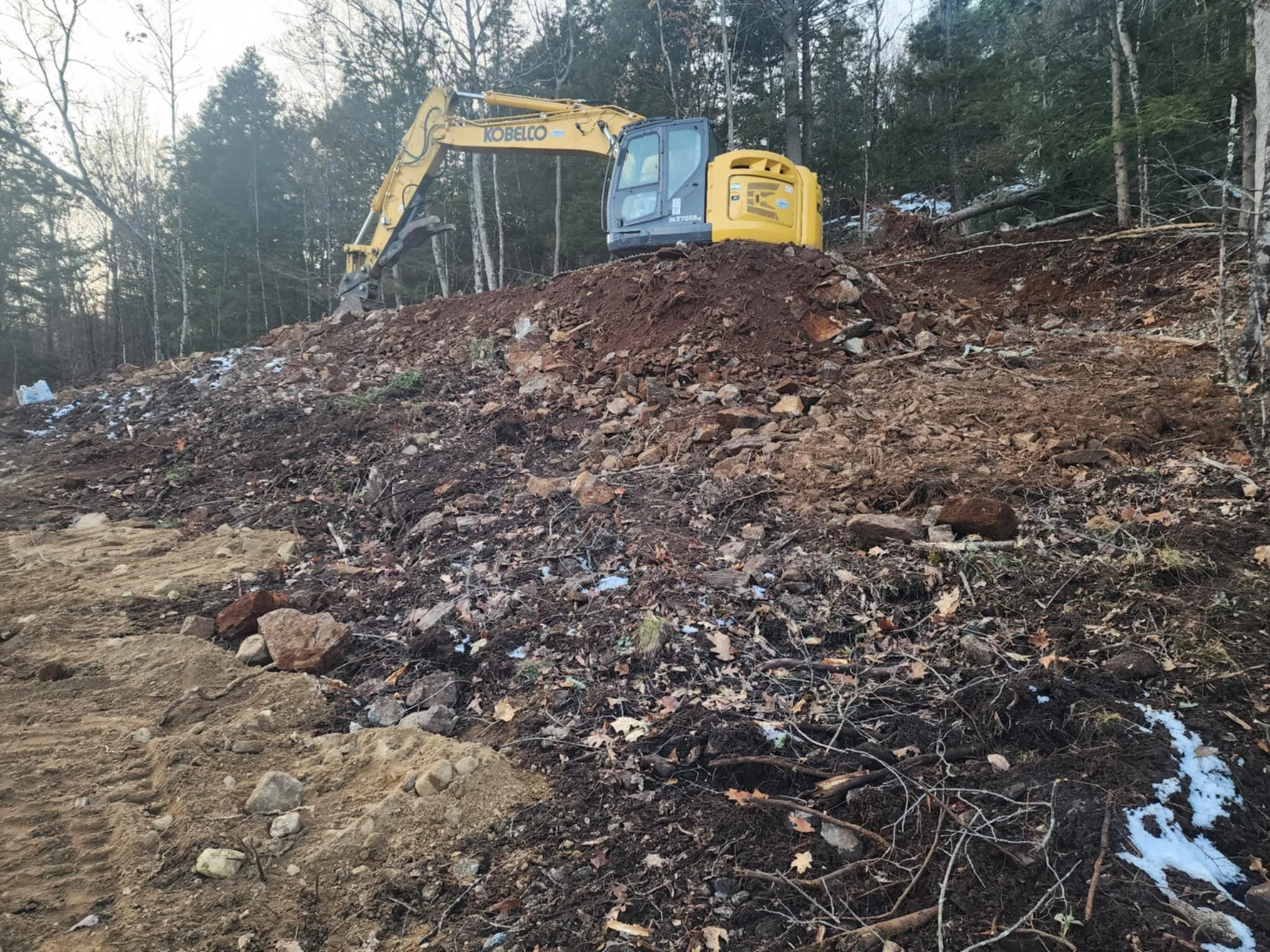 A yellow and black excavator working on a dirt hillside in a forested area with some snow patches on the ground.
