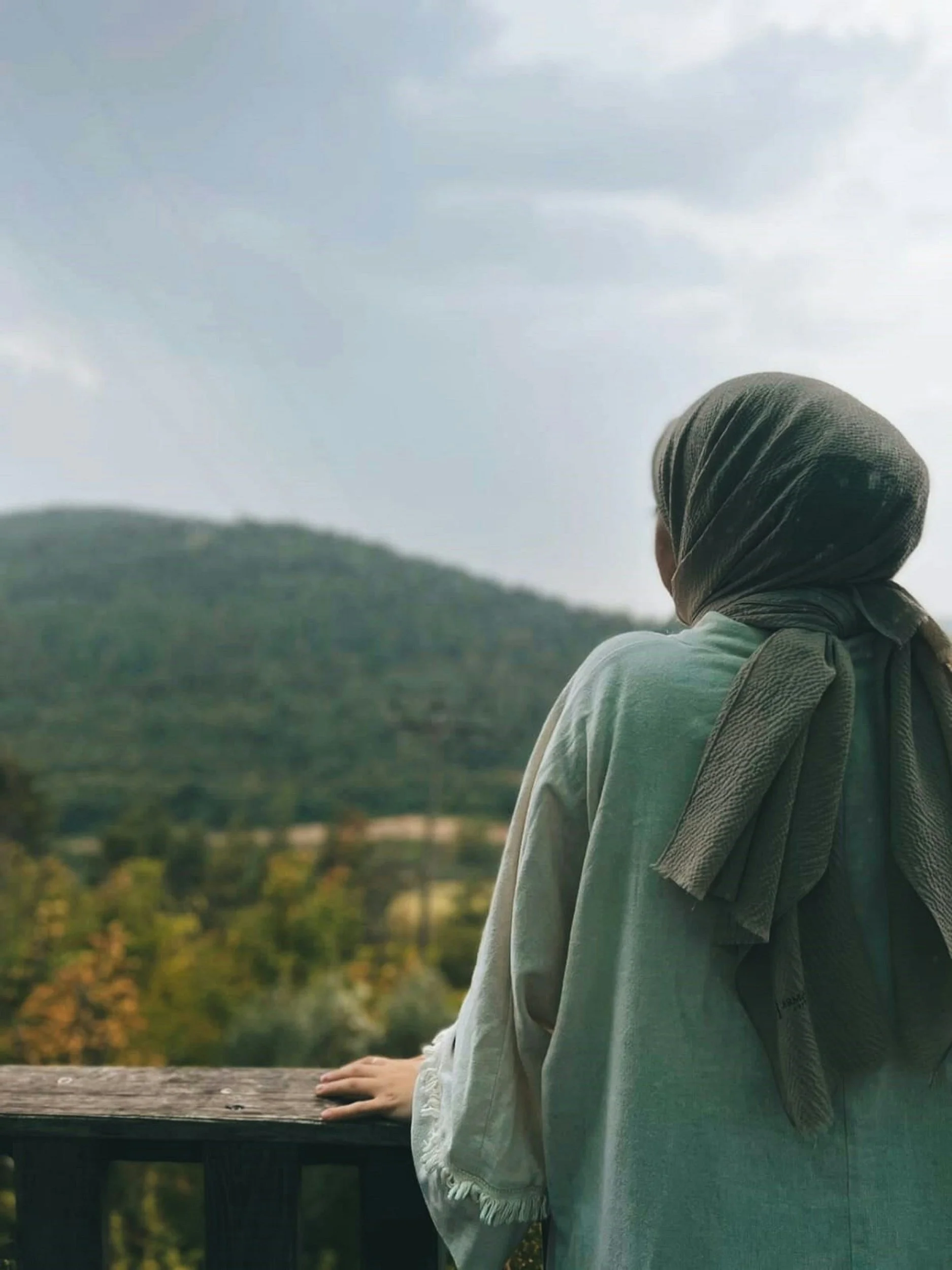 Person wearing a gray headscarf and light gray hoodie, sitting on a wooden railing outdoors with a landscape of trees and hills in the background.