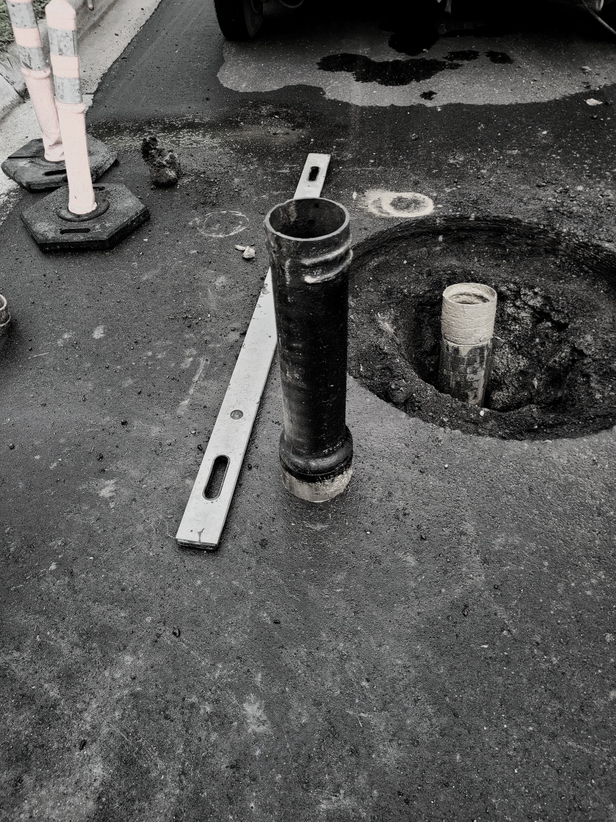Road construction site with a section of asphalt dug out, exposing a pipe sticking out of the ground. A level tool is placed nearby, and construction barriers are visible in the background.