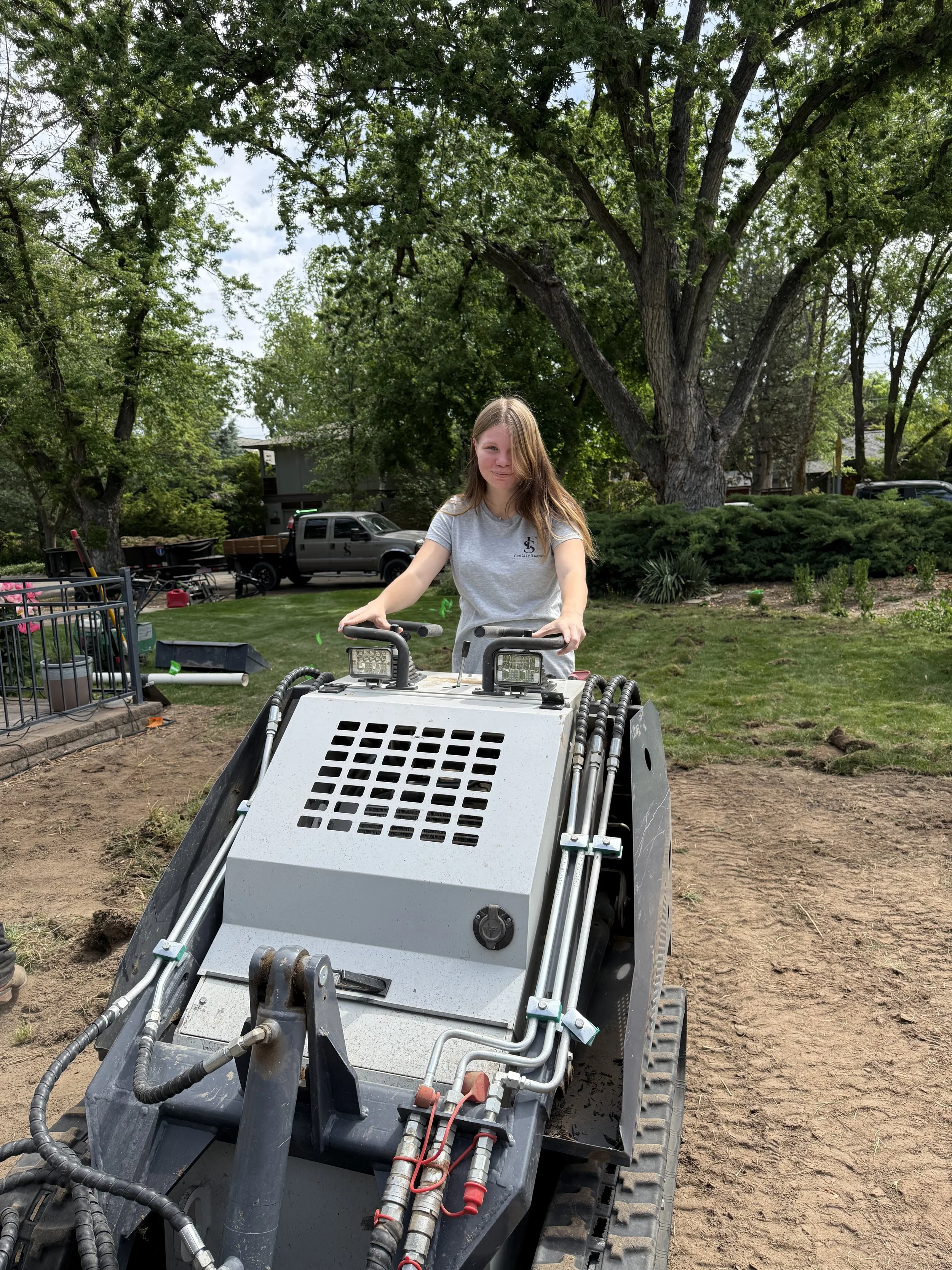A woman with long hair riding a small construction vehicle in a backyard with large trees, shrubs, and parked cars in the background.