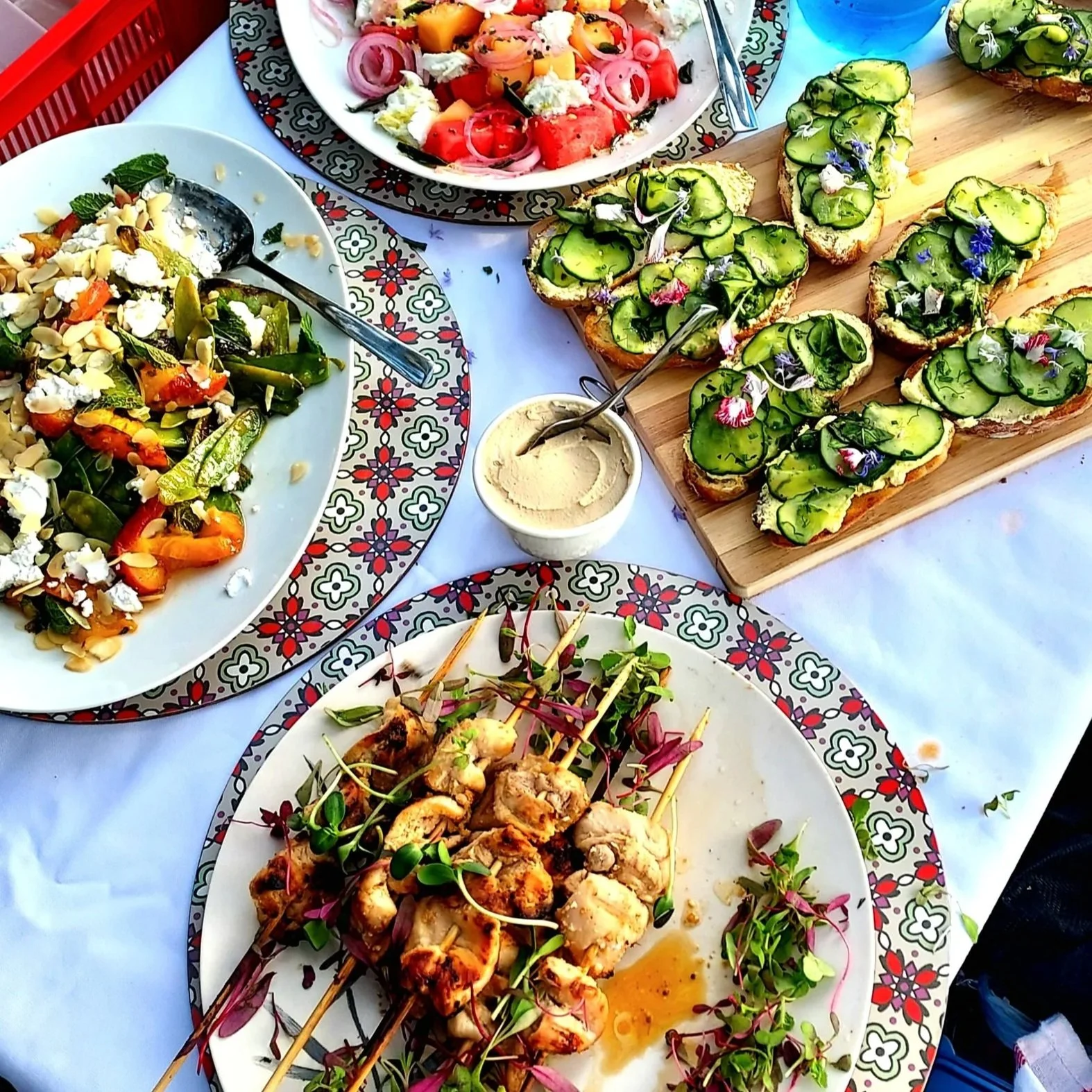 A spread of various colorful dishes on a table, including cucumber-topped toast, grilled chicken skewers, mixed salad, and a bowl of hummus, all arranged on patterned plates and a wooden cutting board.
