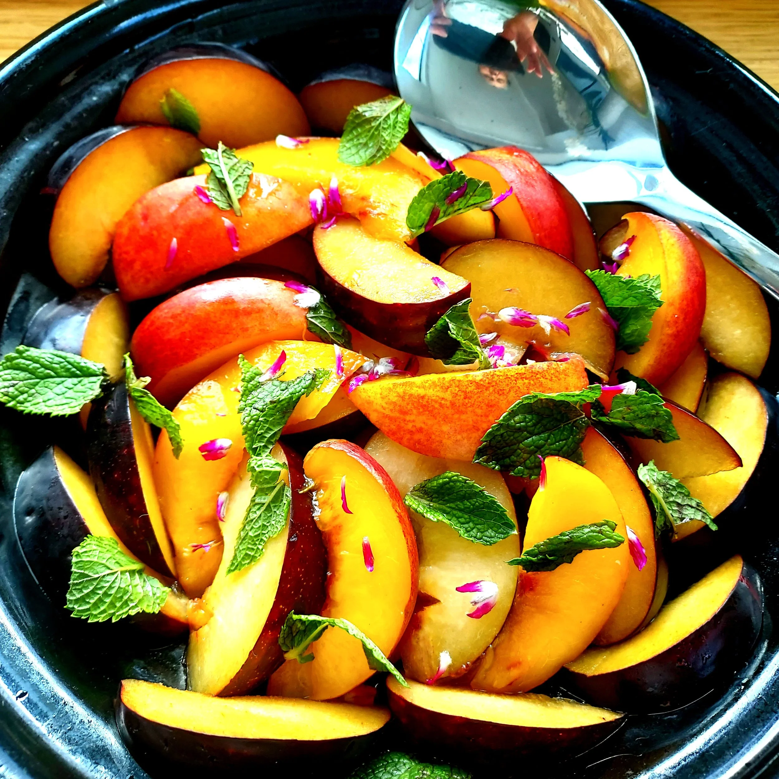 A bowl of sliced peaches garnished with fresh mint leaves and pink flower petals. The peaches are orange and yellow, and some pieces are reflected in a nearby spoon.