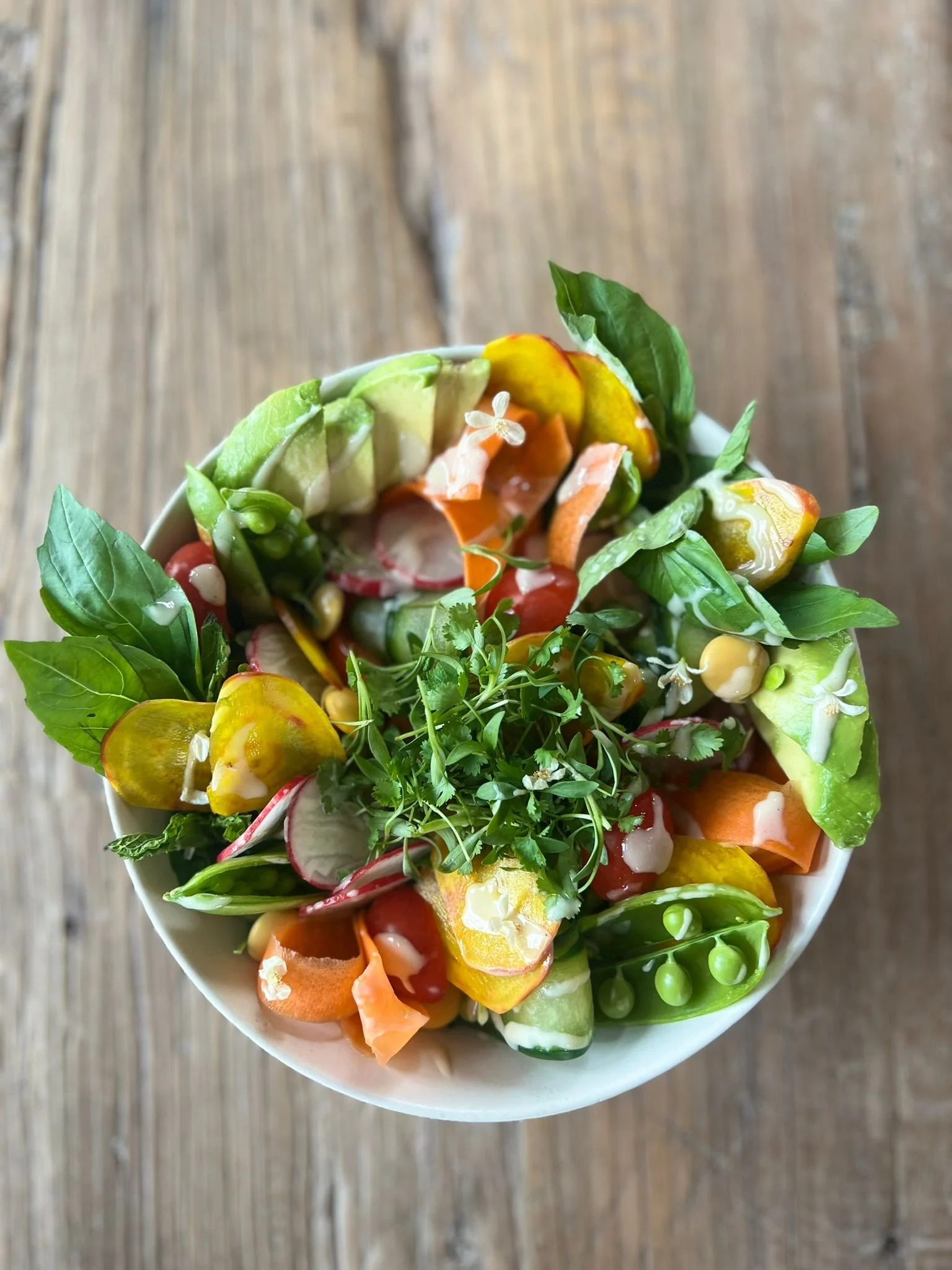 A bowl of fresh salad with various colorful vegetables, including cherry tomatoes, sliced carrots, cucumber, radish, and leafy greens, on a wooden surface.