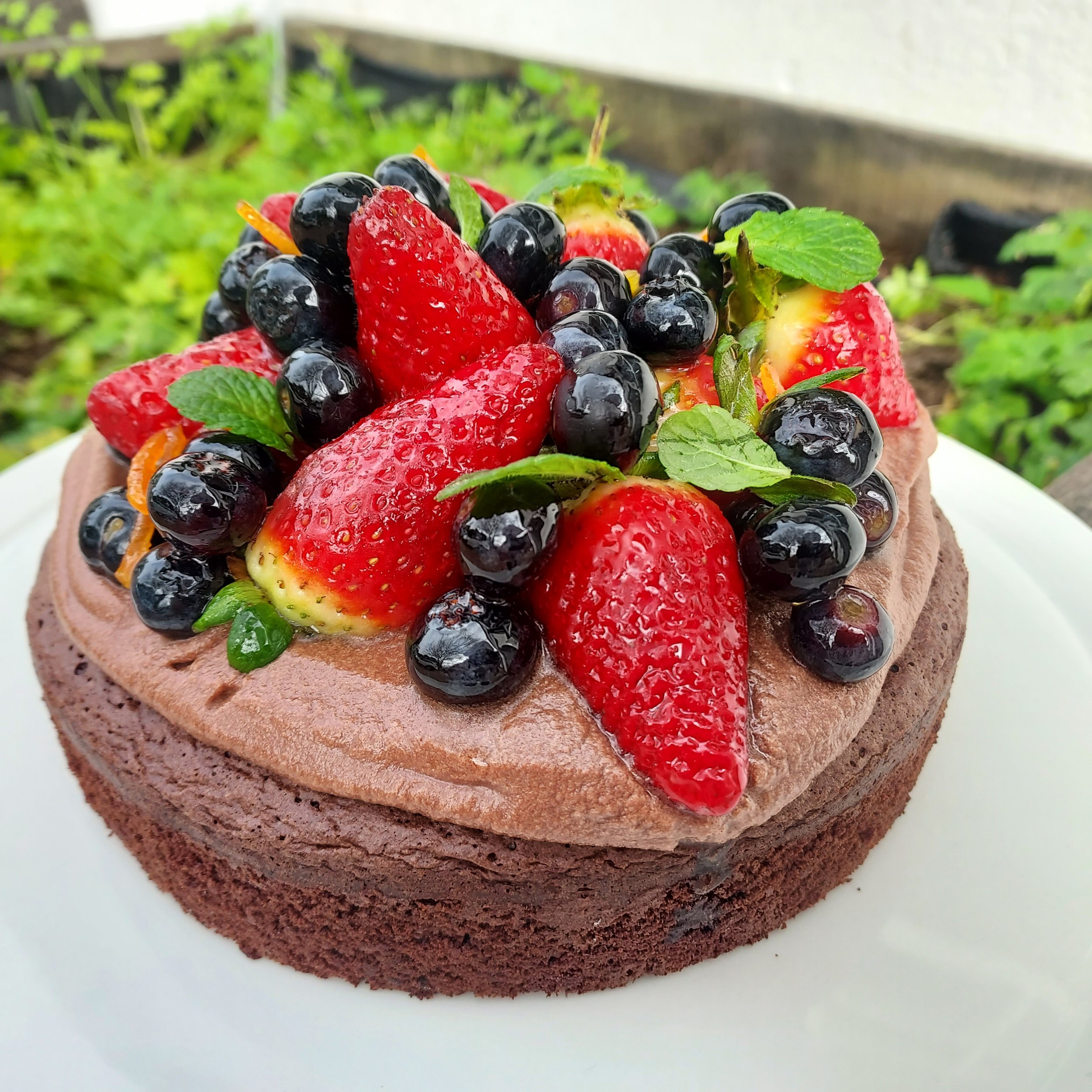 Chocolate cake topped with fresh strawberries, blueberries, mint leaves, and orange zest, on a white surface.