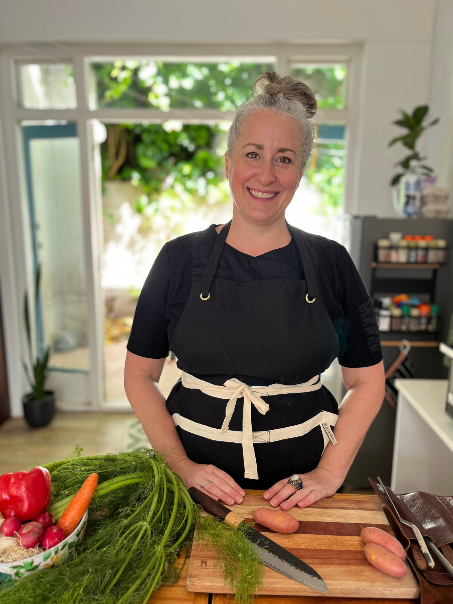 A smiling woman with gray hair tied up, wearing a black apron, standing in a kitchen with a large window, chopping sweet potatoes on a wooden cutting board. Fresh vegetables including radishes, carrots, and herbs are visible on the counter.