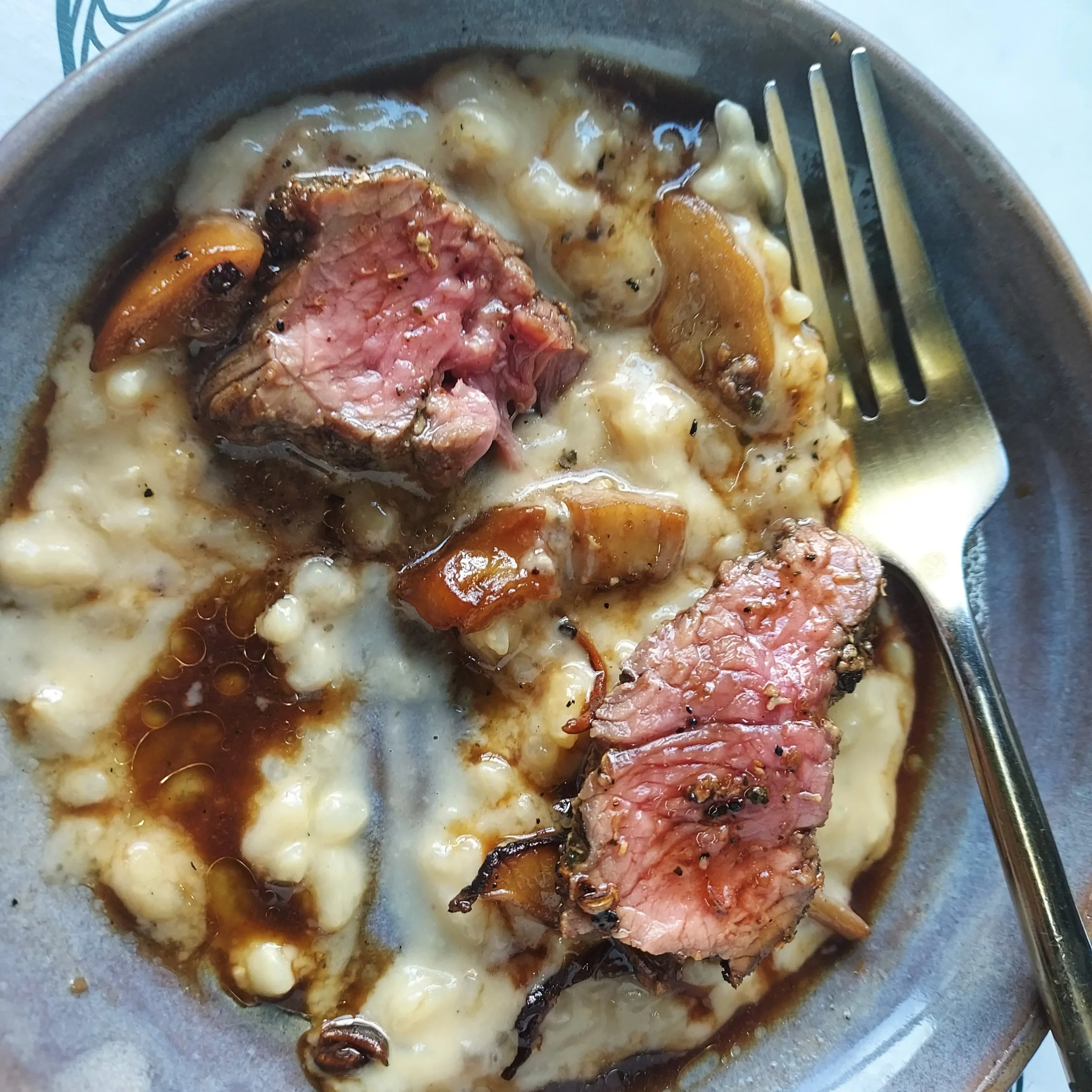 Close-up of a plate with a slice of cooked beef, mashed potatoes, gravy, and mushrooms, with a fork on the side.