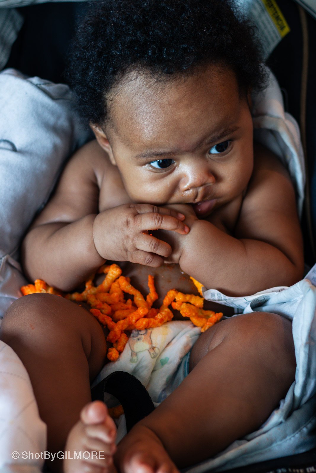 A young child with curly black hair sitting in a car seat with orange snack pieces on their lap, holding one hand near their mouth, looking to the side with wide eyes.