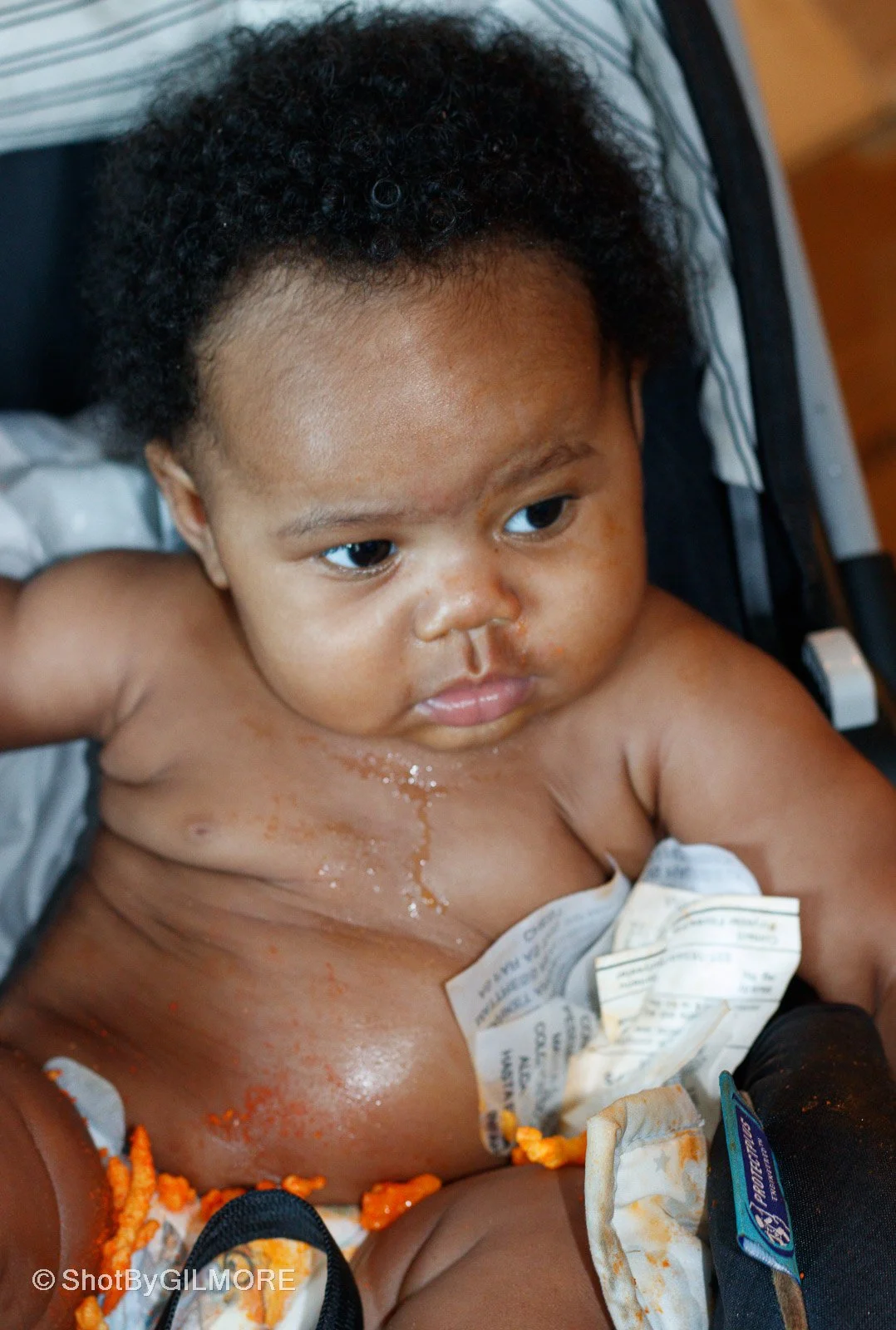 Young child with curly black hair, sitting in a wheelchair, shirtless, with food and some crumpled papers on their chest and lap, looking to the side.