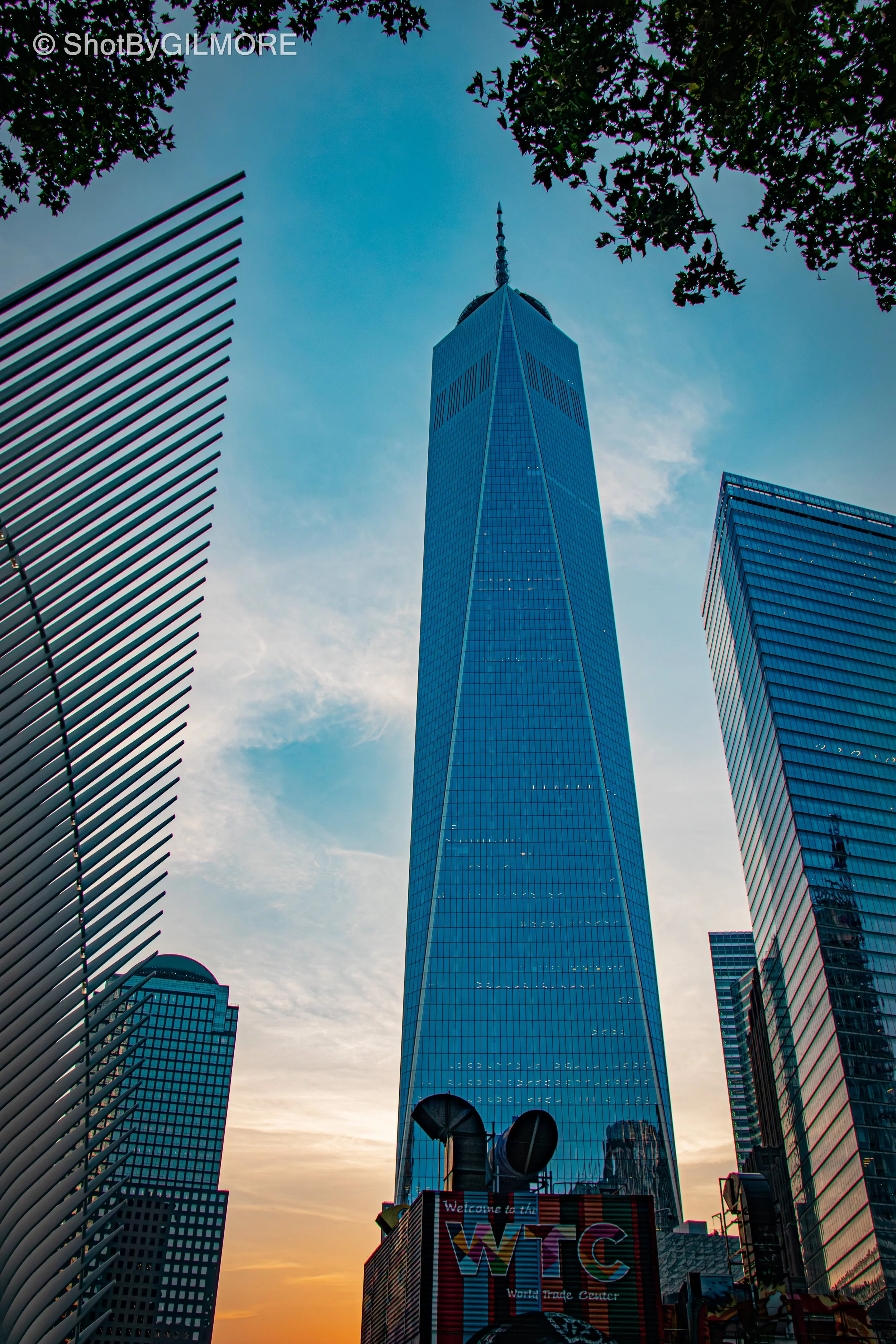 A tall skyscraper with a spire viewed from the street level at sunset, surrounded by other tall buildings and trees.