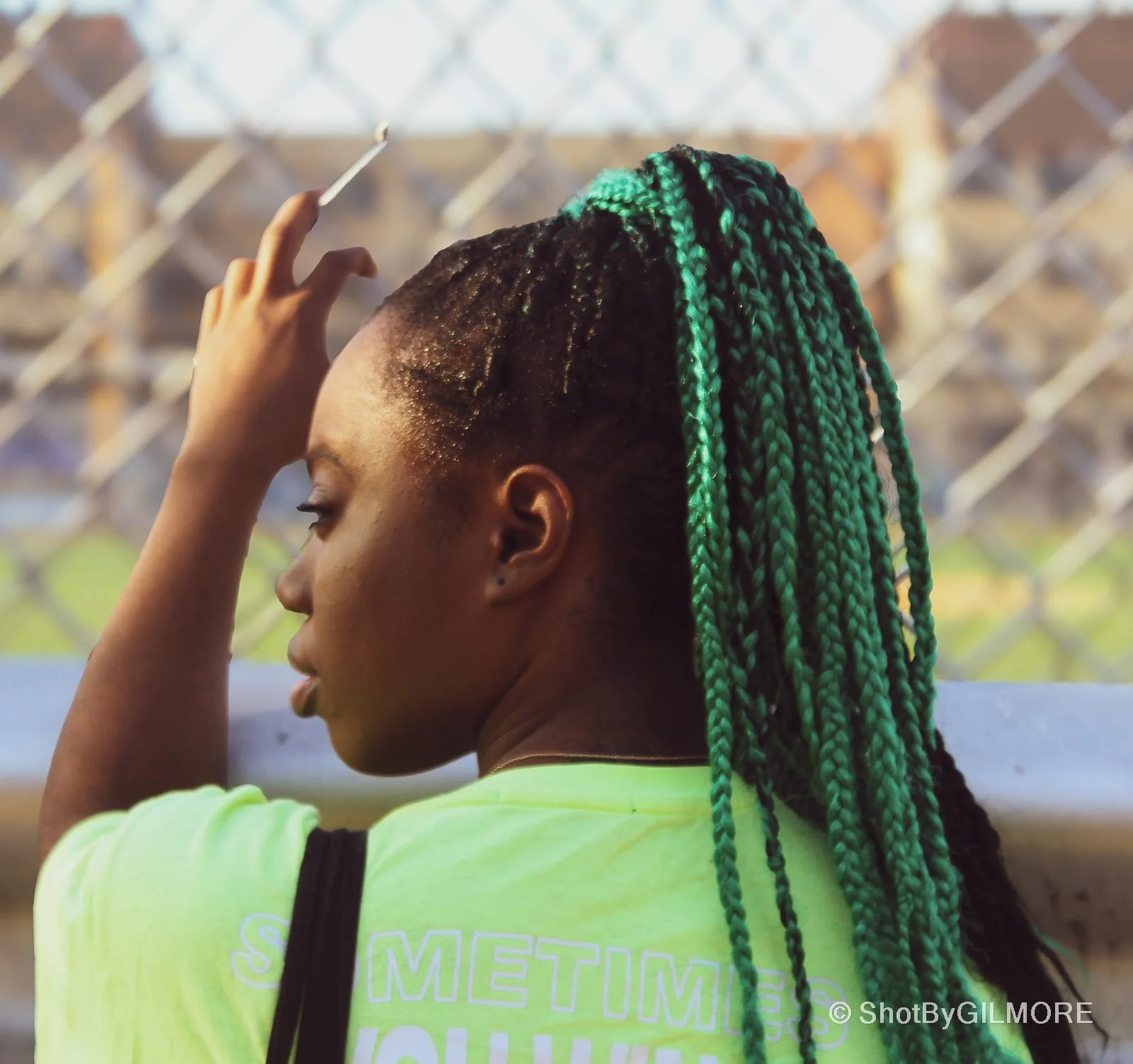 A woman with long green braided hair, resting her head against a fence, gazing down, wearing a neon green shirt.