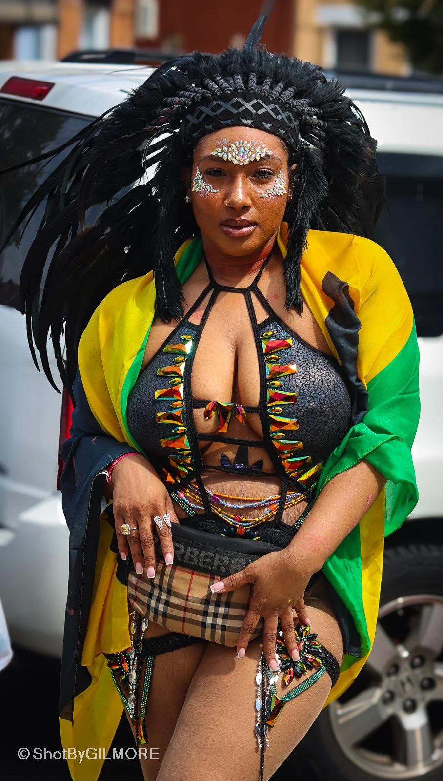 Woman dressed in colorful carnival costume with feathers, face gems, and revealing clothing, standing outdoors near cars.