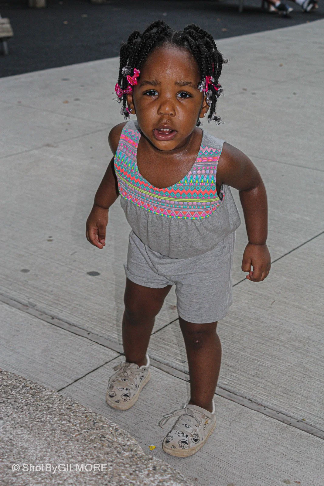 A young girl with curly hair in braids decorated with pink and silver beads, standing on a sidewalk and looking up with a distressed or curious expression.