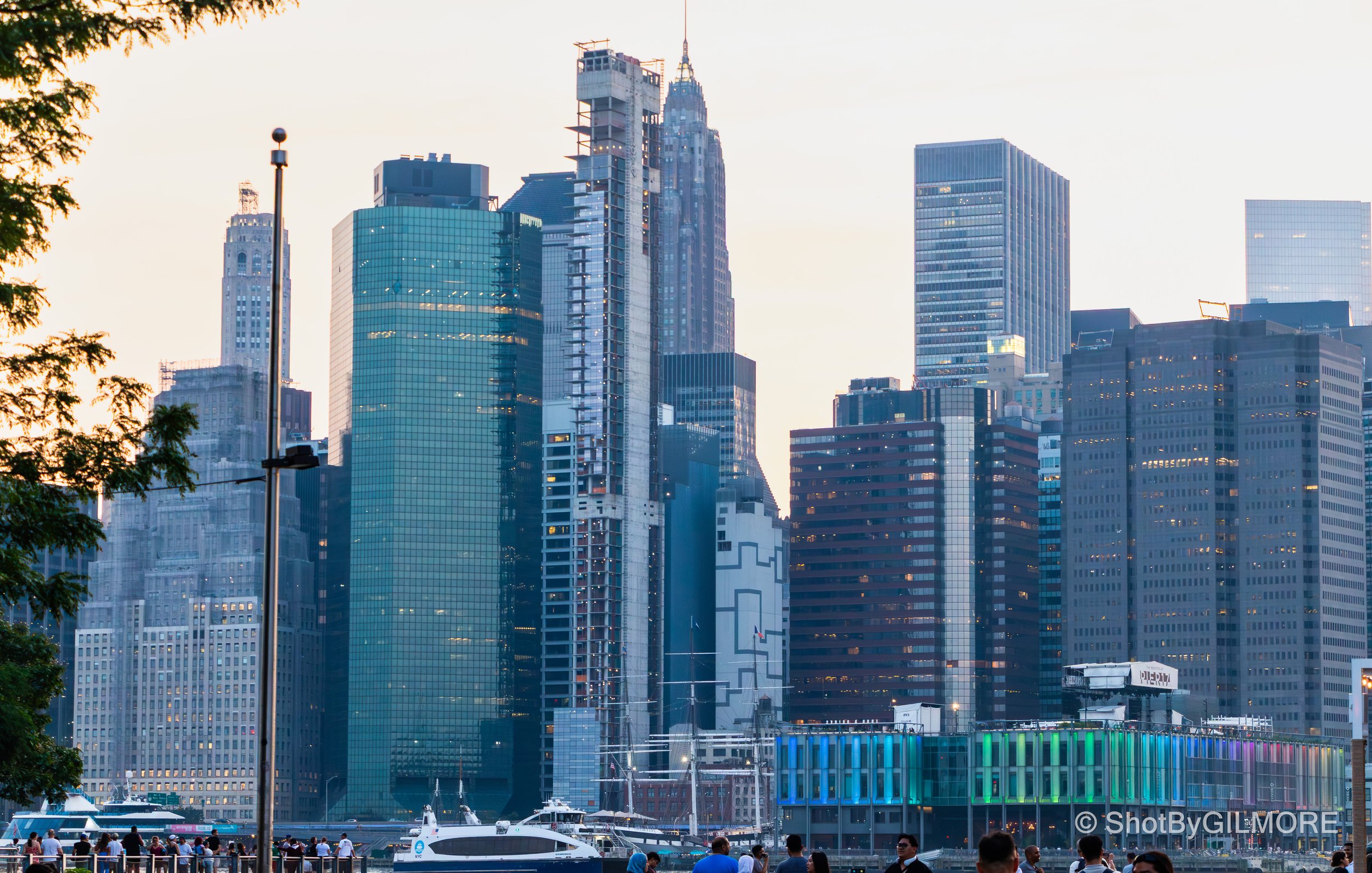 View of a city skyline with tall skyscrapers, some under construction, and boats docked along the waterfront, with people walking in the foreground.