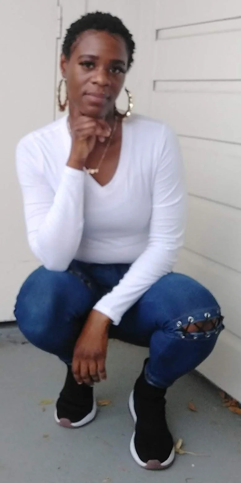 A woman sitting on her knees in front of a white wall, wearing a white long-sleeve shirt, ripped jeans, and black sneakers.