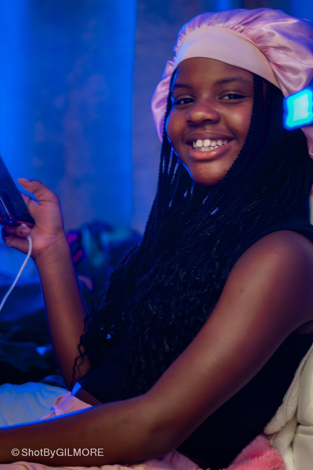 A young girl with braided hair smiling while holding a phone, wearing a pink satin bonnet, in a dimly lit blue environment.