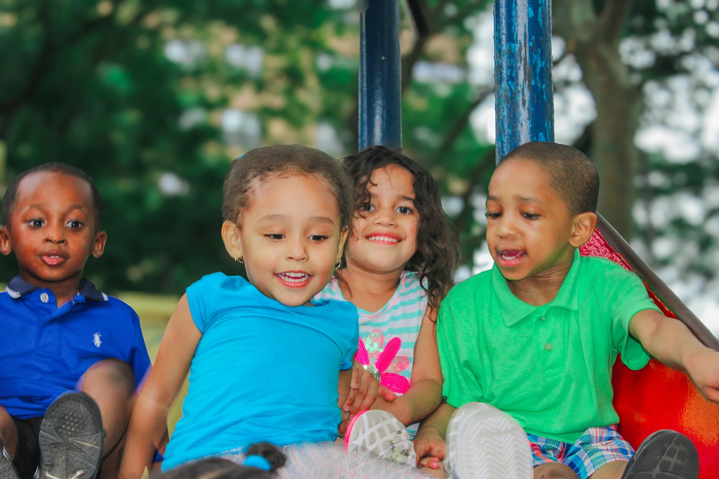 Four children sitting together outdoors in a park on a swing set, smiling and enjoying themselves.