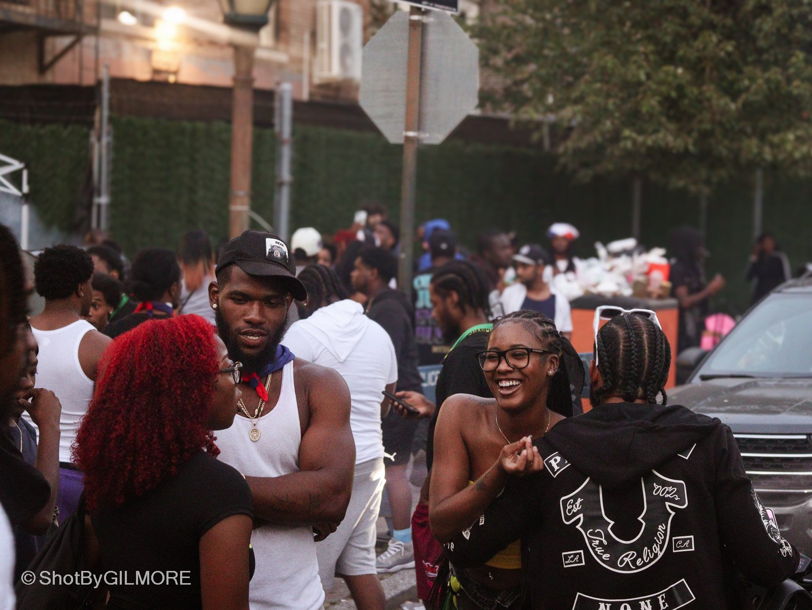 A group of people socializing outdoors, some wearing casual clothing, with a few smiling and engaging in conversations. There are trees, a car, and a street sign in the background.