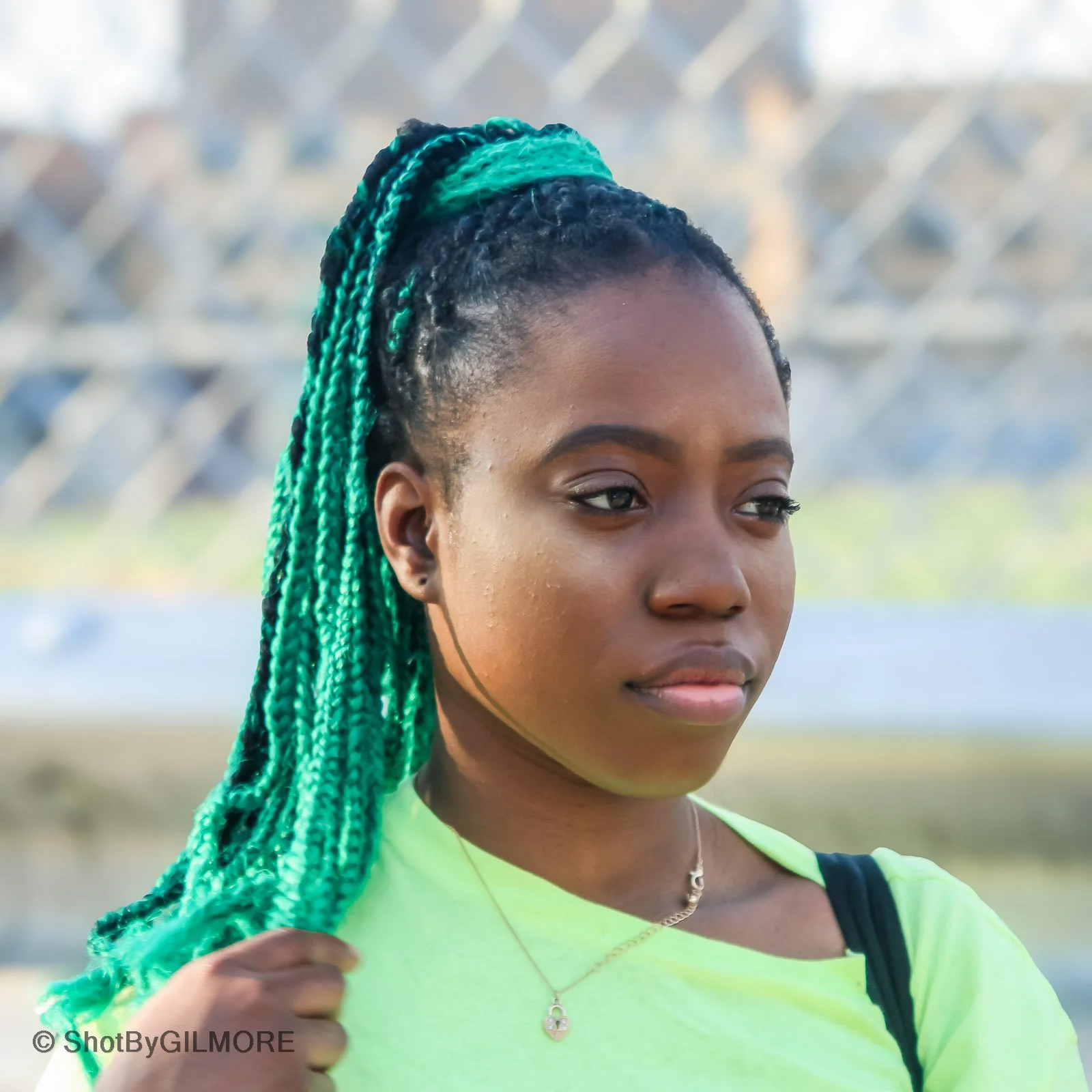 A young woman with green and black braids, wearing a neon yellow shirt and a delicate necklace, stands outdoors with a blurred background of a chain-link fence and greenery.