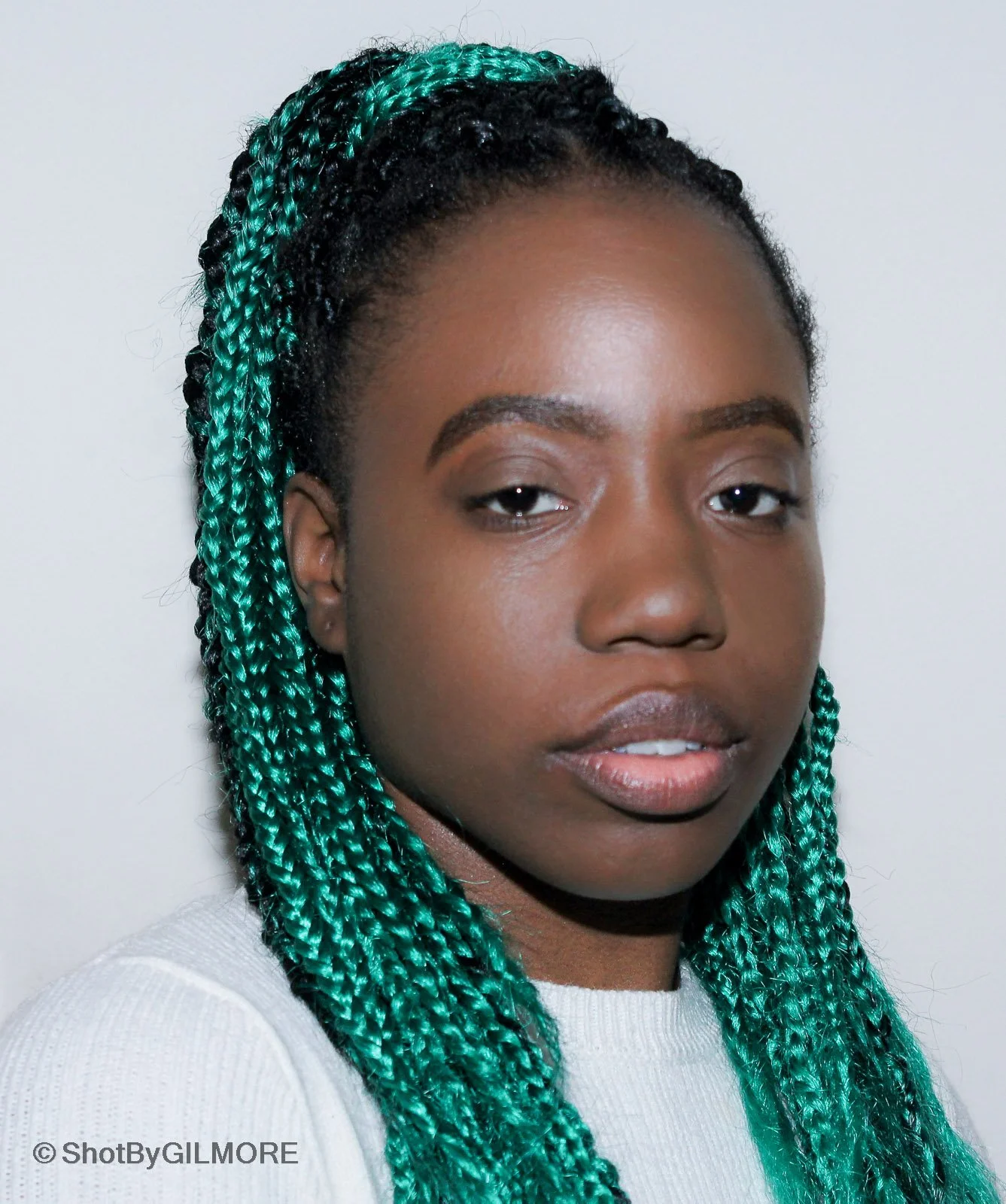 A young woman with dark skin and teal braided hair, wearing a light-colored top, looking at the camera with a neutral expression, against a plain white background.