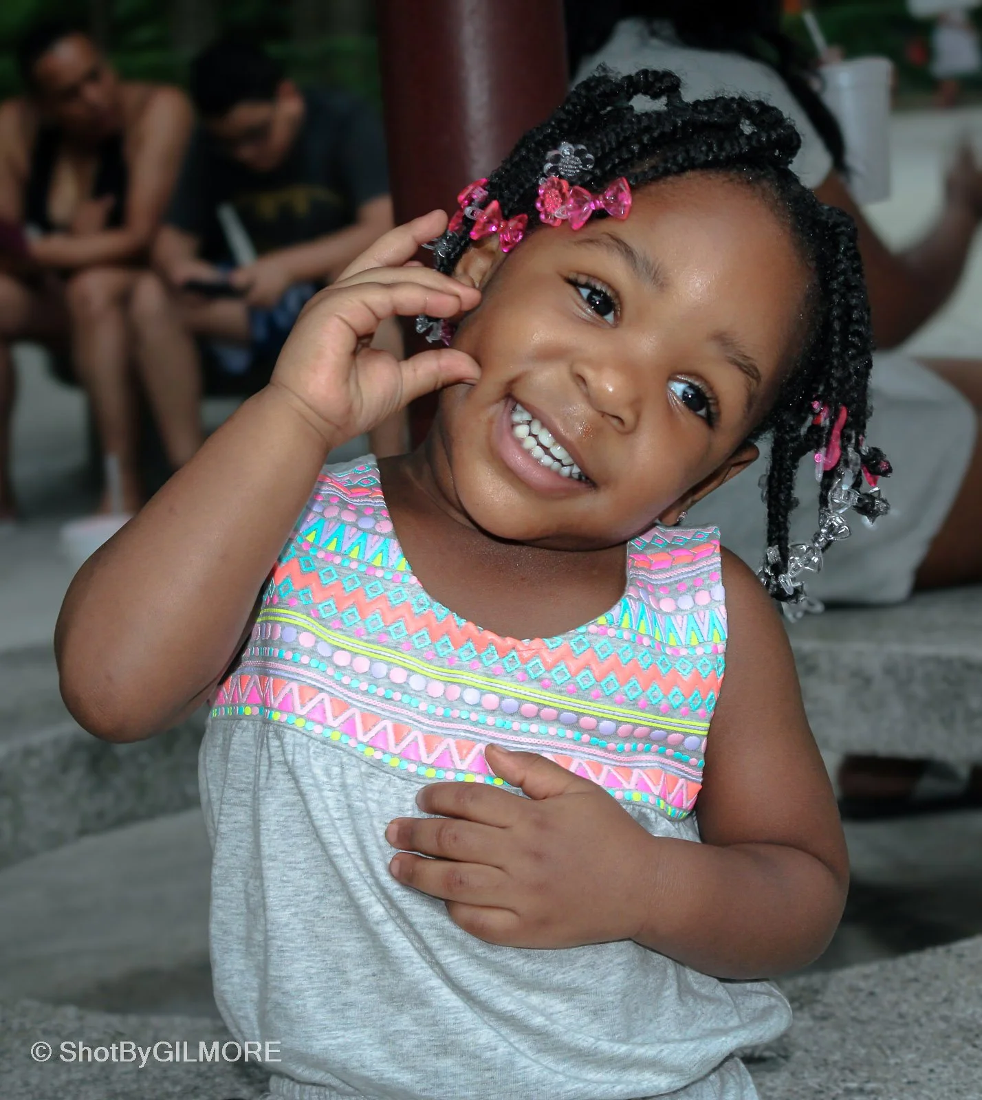 A young girl smiling happily, with her hand touching her cheek, wearing a colorful sleeveless top with pink, blue, and yellow patterns, and hair styled in braids with pink and clear hair accessories.
