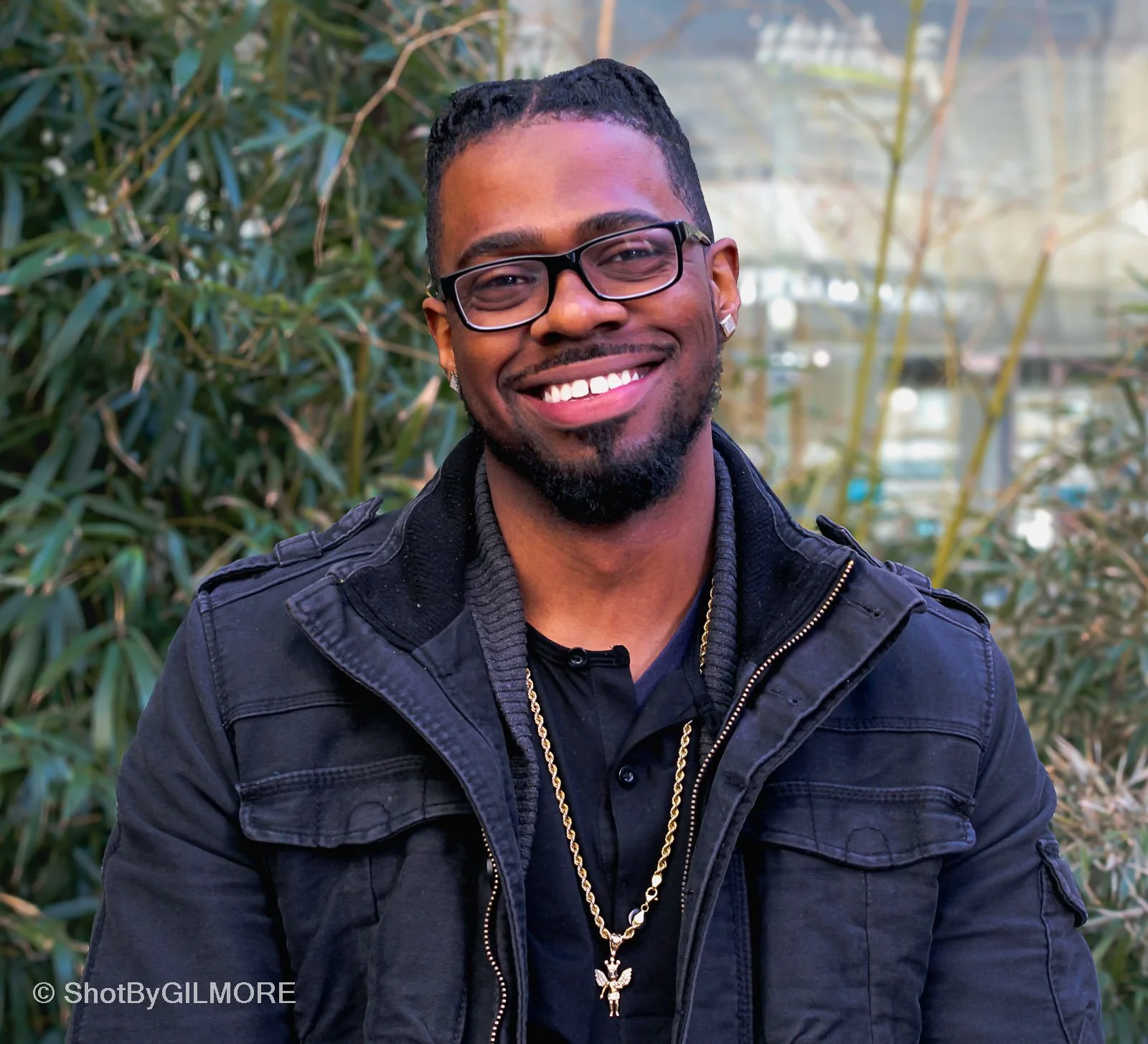 A smiling man with glasses and facial hair, wearing a black jacket and gold jewelry, standing in front of green foliage.