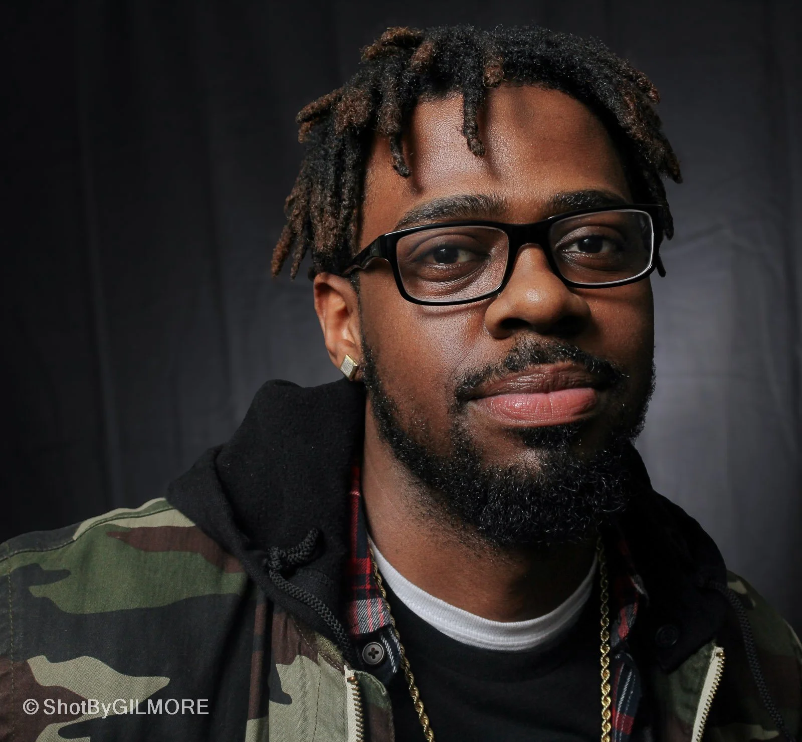 A young man with glasses, earrings, and facial hair, wearing a camouflage jacket over a plaid shirt and a gold chain, posing against a black background.