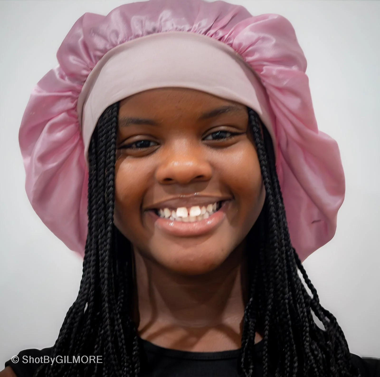 Close-up of a young girl smiling with black braided hair, wearing a pink satin bonnet and a beige headband, against a plain light background.