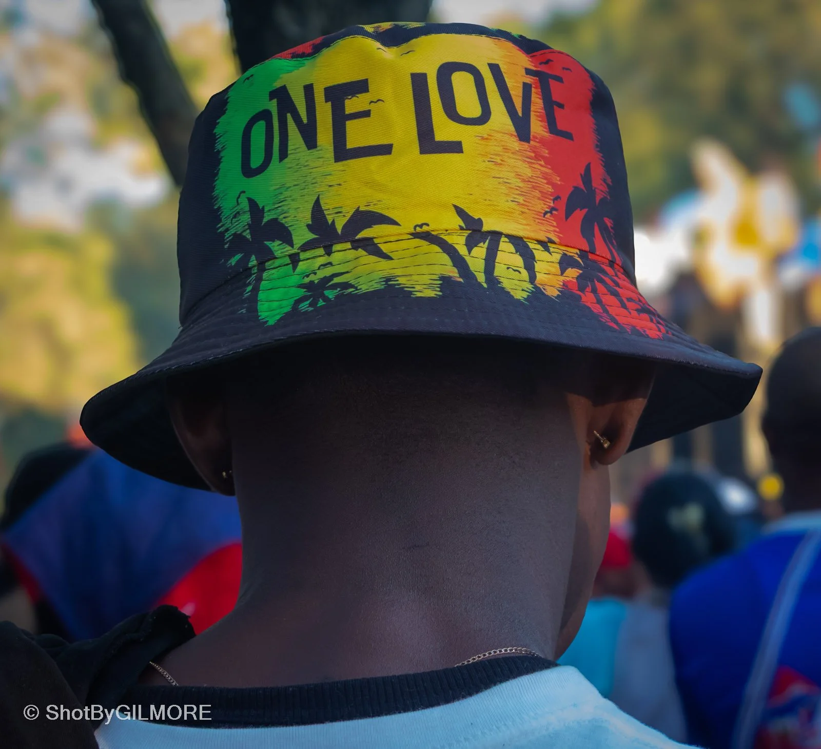 Person wearing a colorful bucket hat with the words 'ONE LOVE' and tropical palm tree graphics, facing away in an outdoor setting with trees and crowd in background.