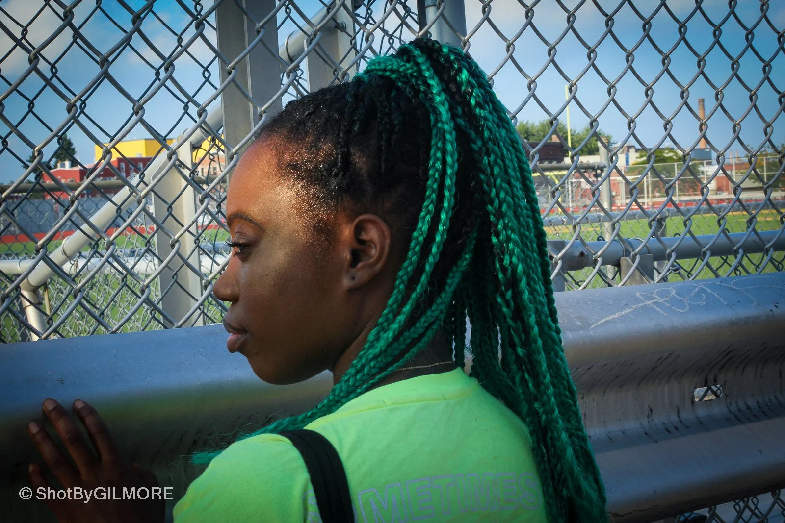 A young woman with green and black braids leaning on a baseball field fence, looking down, with a baseball field and sky in the background.