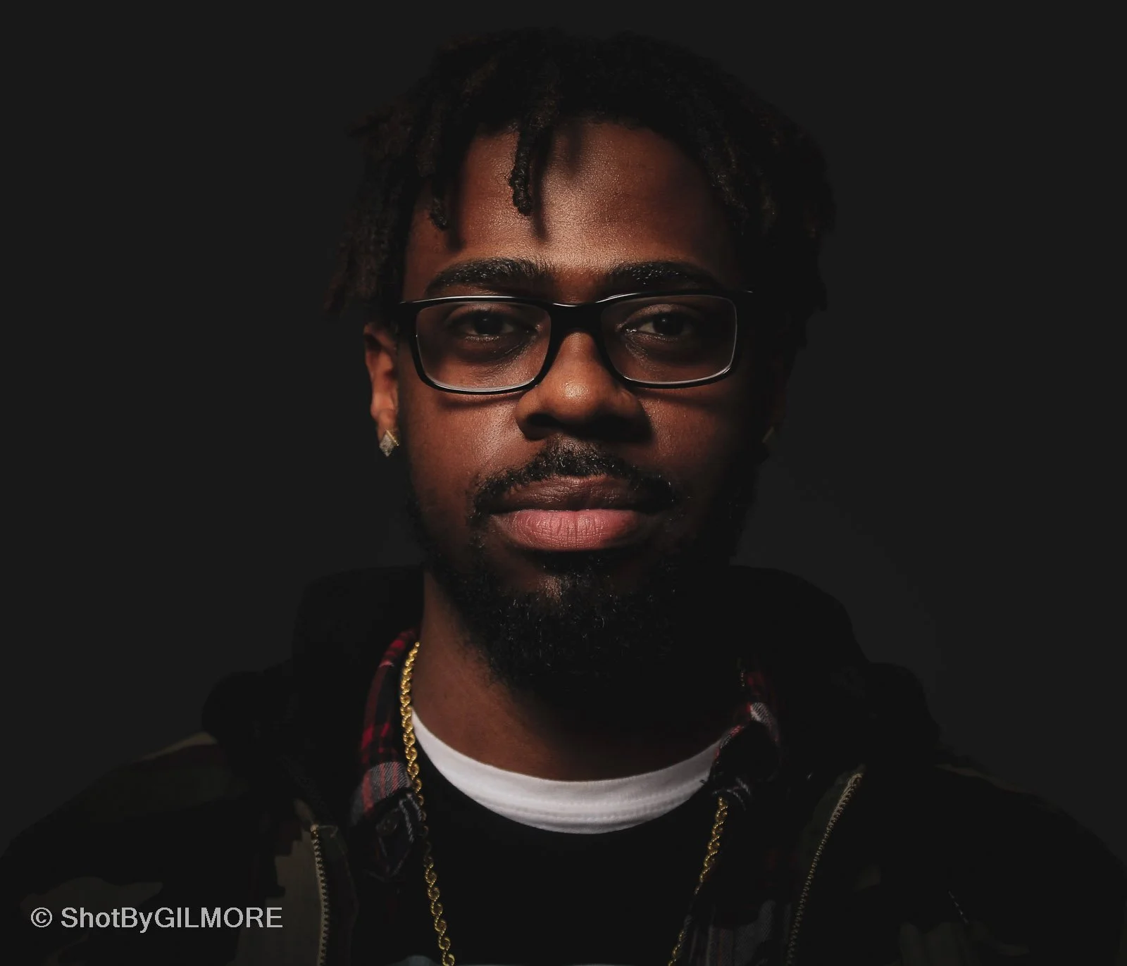 Portrait of a young man with glasses, a beard, earrings, and dreadlocks, wearing a hoodie and gold chain against a dark background.