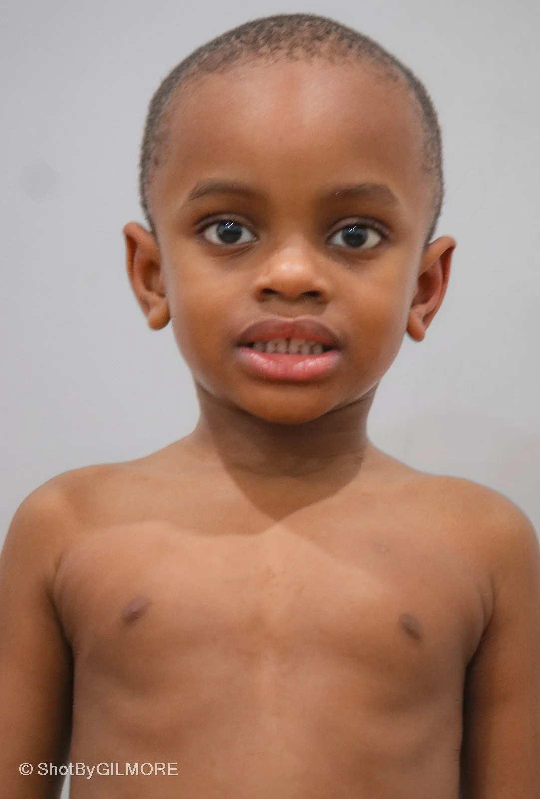 Close-up of a young African American boy with short hair, shirtless, looking at the camera against a plain background.