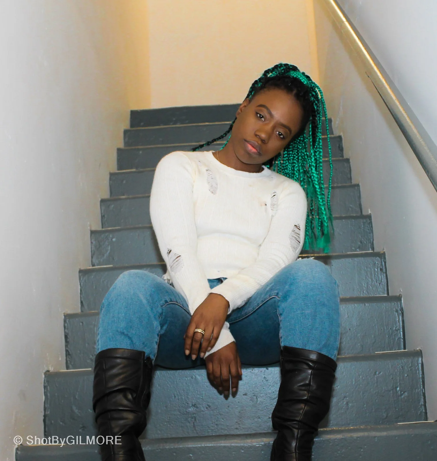 A young woman with green braided hair sitting on gray stairs, wearing a white distressed sweater, blue jeans, and black boots, with a neutral expression.