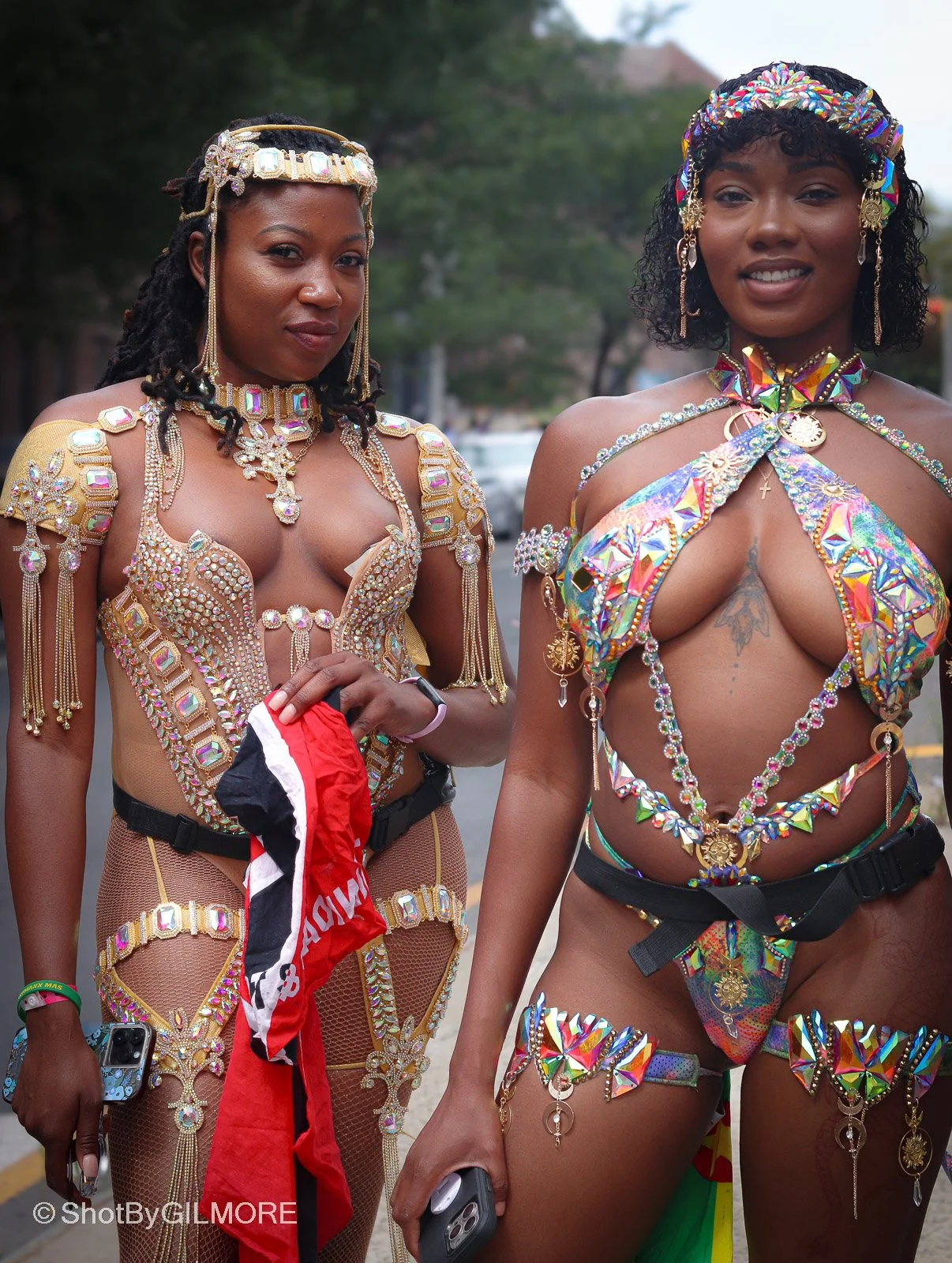 Two women dressed in colorful, elaborate costumes with rhinestones, beads, and jewelry, standing outdoors at a festival or event.