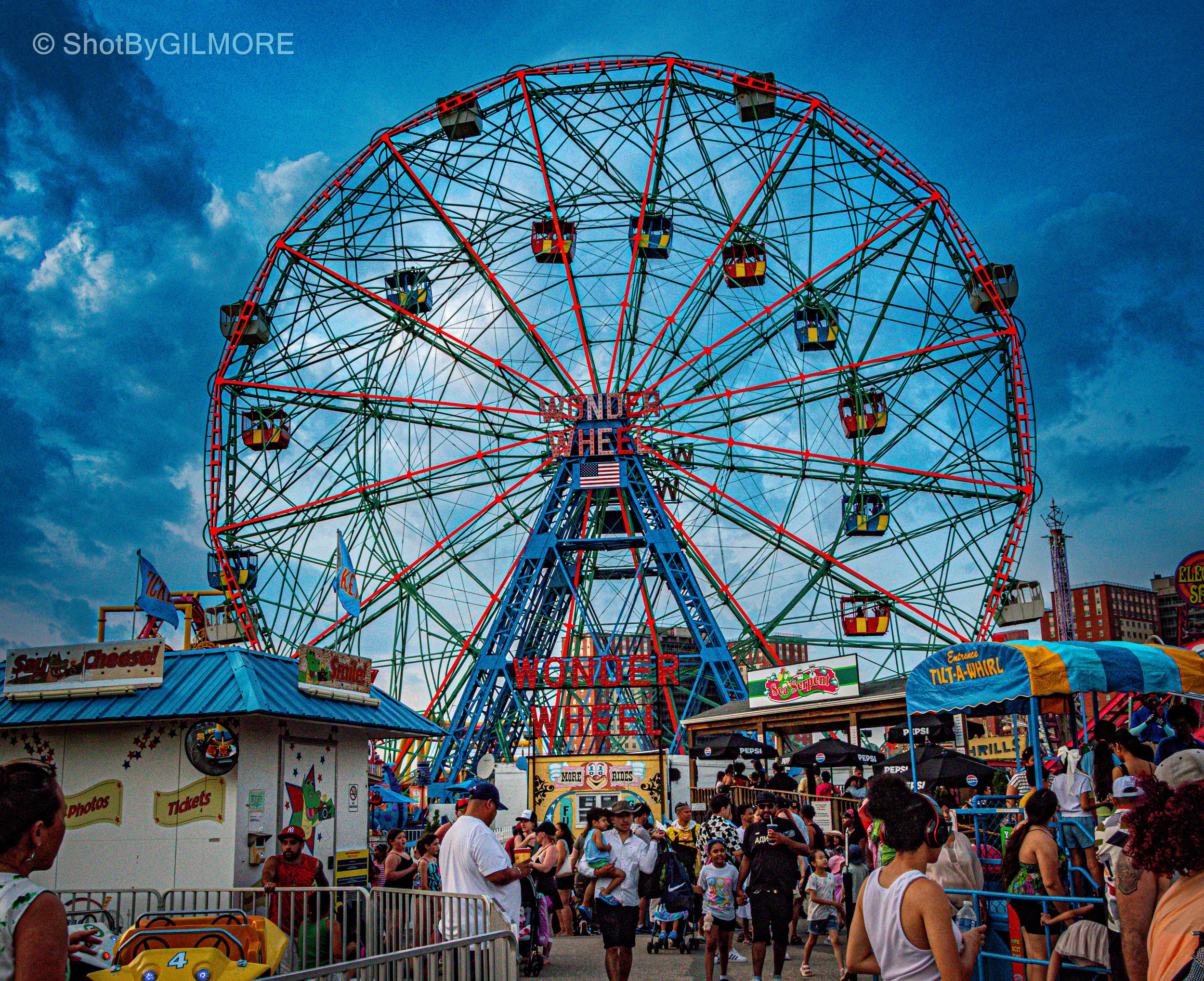A large red and green Ferris wheel in a carnival, with people waiting in line and walking around in the foreground, vendor booths, and a cloudy sky in the background.