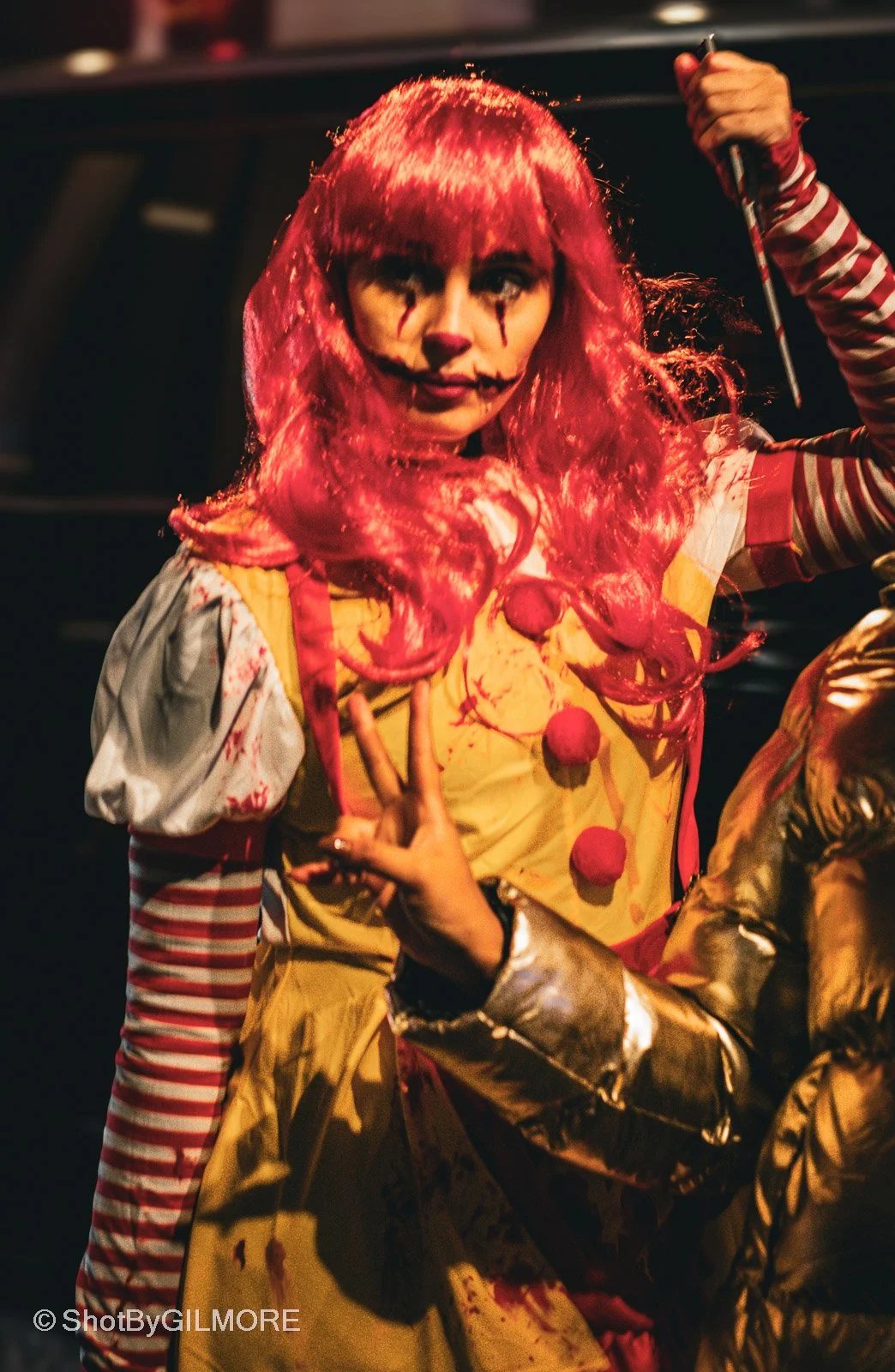 A woman dressed as a creepy clown with red wig, painted face, and clown costume, making a peace sign with her fingers.