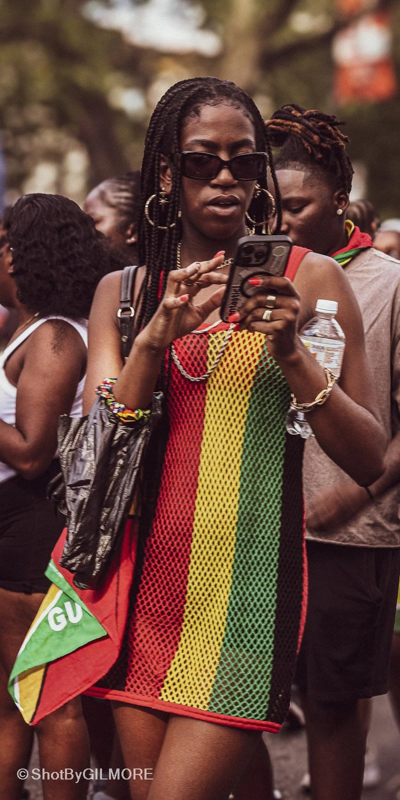 A woman with large earrings and sunglasses looking at her phone, wearing a mesh dress with red, yellow, green, and black vertical stripes, surrounded by other people at an outdoor event.