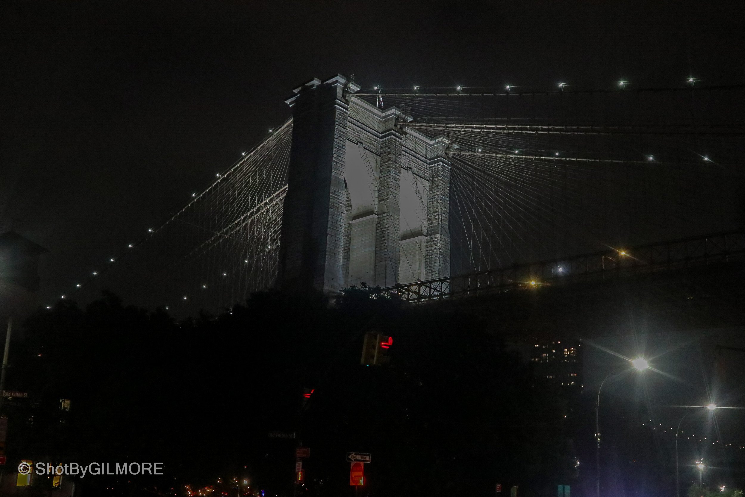 Nighttime view of the Brooklyn Bridge illuminated, with its cables and towers visible against the dark sky, and city lights reflecting on the river below.