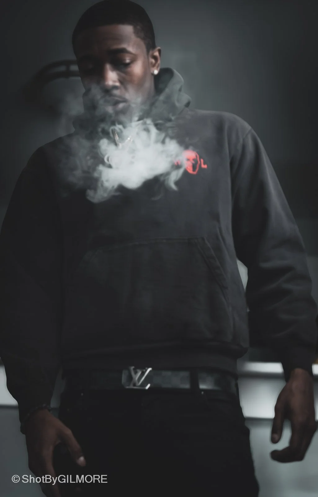 A young man with dark skin wearing a black hoodie and black pants, with a Louis Vuitton belt, is blowing out a cloud of smoke or vapor.