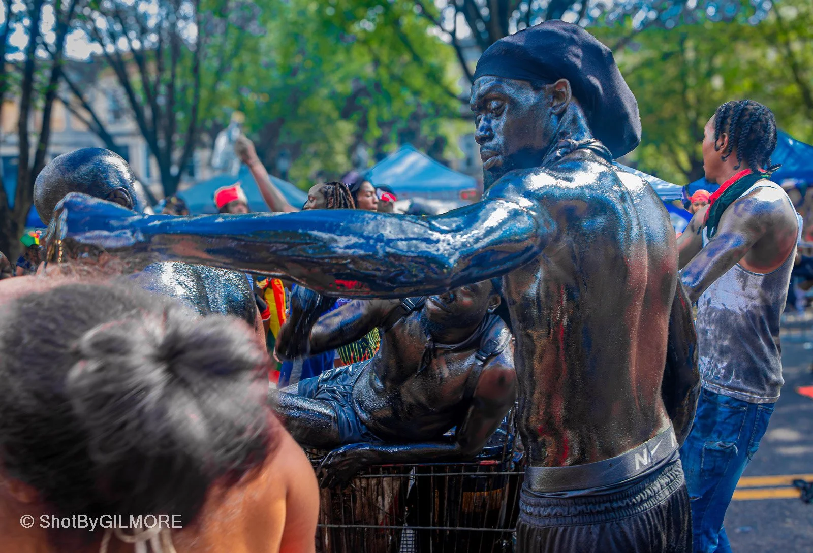 People celebrating at a festival, some covered in black mud or paint, outdoors with tents and trees in the background.