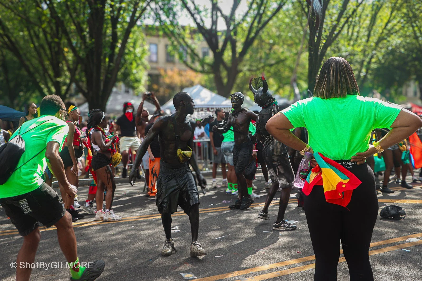 People dancing and celebrating on a city street during a festival or parade, with trees and tents in the background.