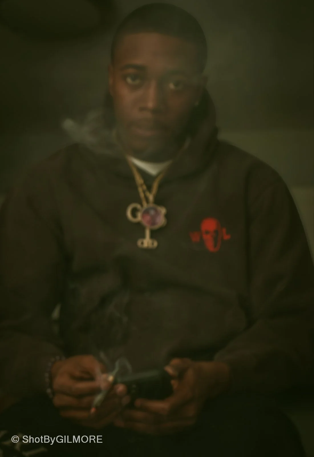 A young man with short hair and dark skin sitting, wearing a black hoodie with a red face logo, accessorized with multiple gold chains, one with a large pendant, and holding a smoking cigarette and a smartphone.