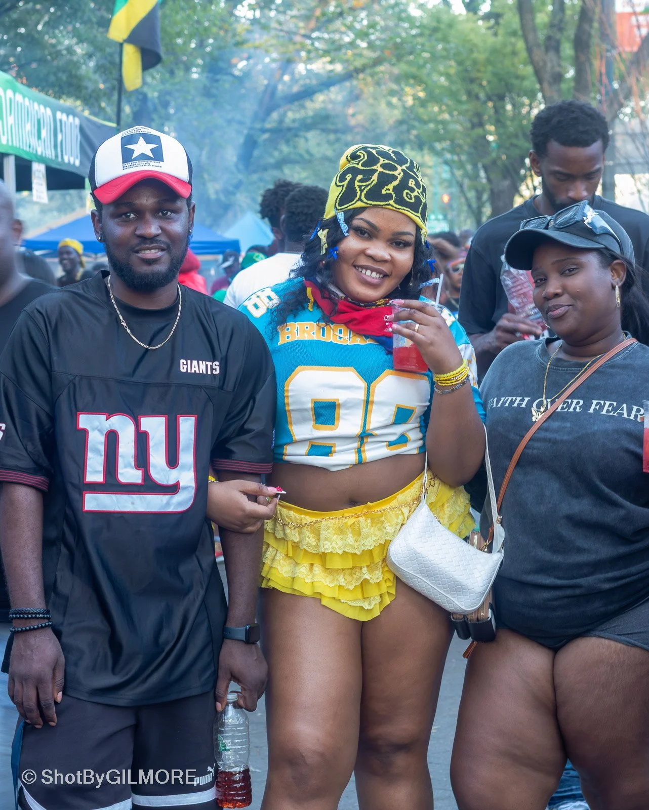 Three people standing outdoors at a festival or event, dressed casually, with the middle person wearing a colorful football jersey, yellow ruffled shorts, and holding a drink, smiling at the camera.