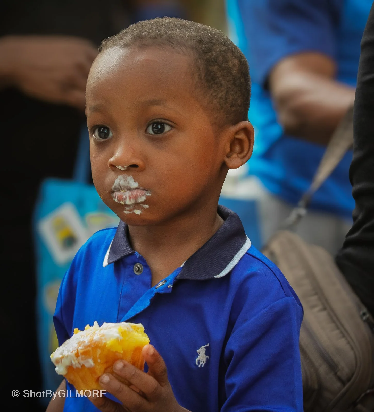 Young boy with short hair and dark skin holding a cupcake with yellow icing, some leftover frosting on his face, wearing a blue polo shirt with a small white logo, in a crowded setting.